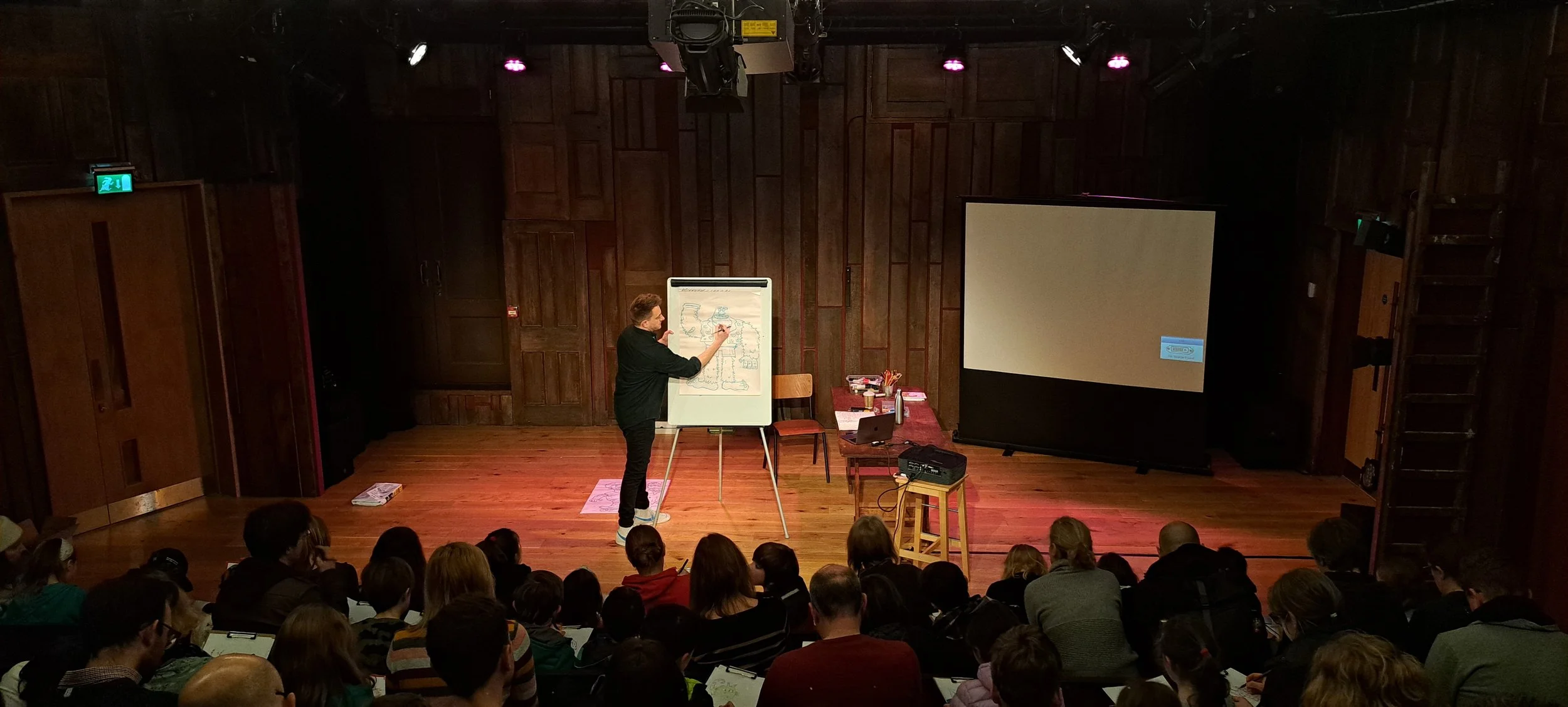 A man giving a presentation to an audience in a wooden-walled auditorium. He is drawing on a whiteboard while standing next to a table with books, papers, and a laptop. There is a large projection screen on the right side and people are seated facing him.