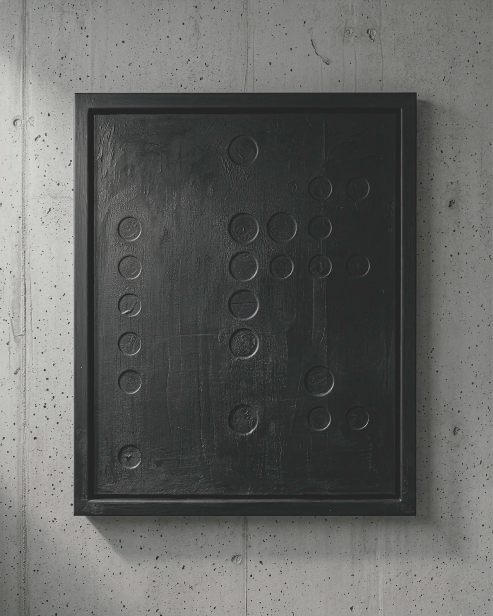 Framed black textured art piece with circular indentations on a concrete wall.