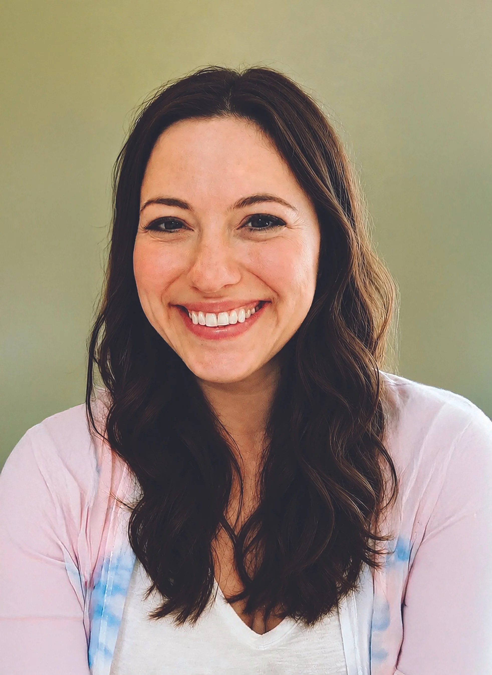 Close-up of a young woman with long wavy brown hair, smiling, wearing a white top and a light pink jacket with blue accents, against a neutral green background.