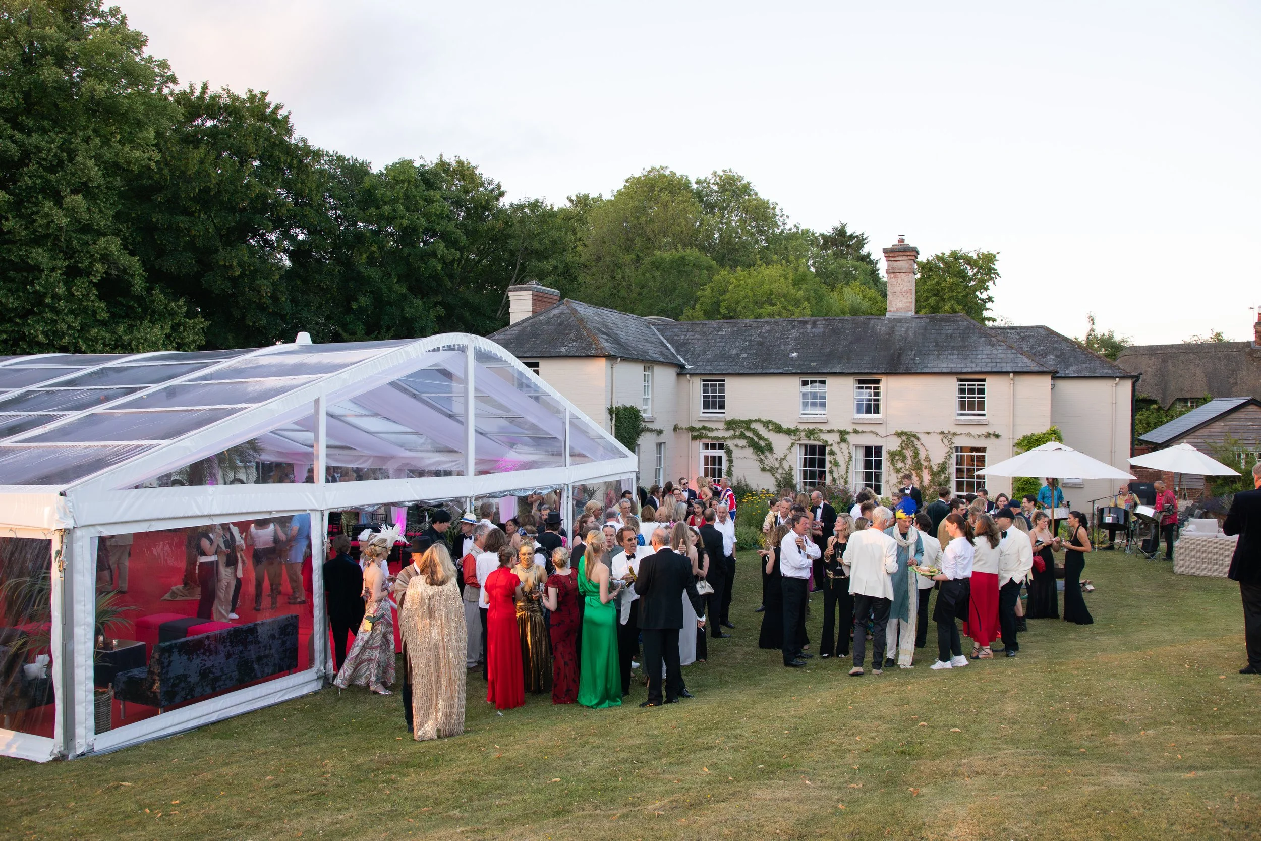 A large outdoor gathering on a lawn in front of a white house with ivy, during daytime. People are dressed in formal attire, with women in colorful dresses and men in tuxedos. A clear marquee tent is visible in the foreground, and the scene is surrou