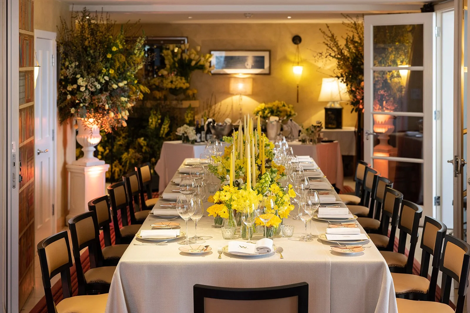 An elegantly decorated dining room set for a formal event, featuring a long table with a white tablecloth, yellow floral centerpieces, tall candles, and place settings, with large floral arrangements and warm lighting in the background.