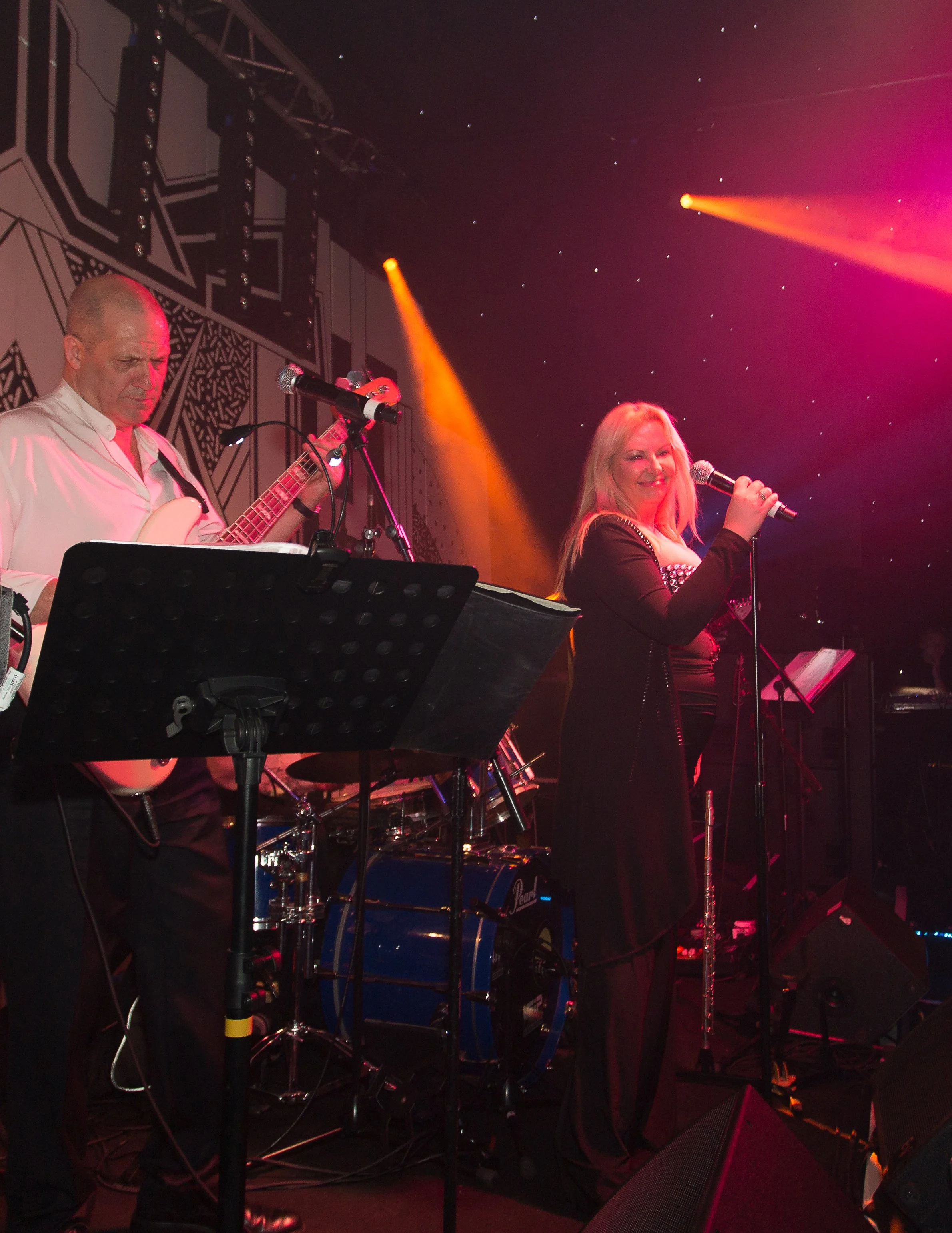 A band of three performers on stage with musical instruments and microphones, illuminated by colorful stage lights, including an orange beam and pink lighting, with a dark background featuring small star-like lights.