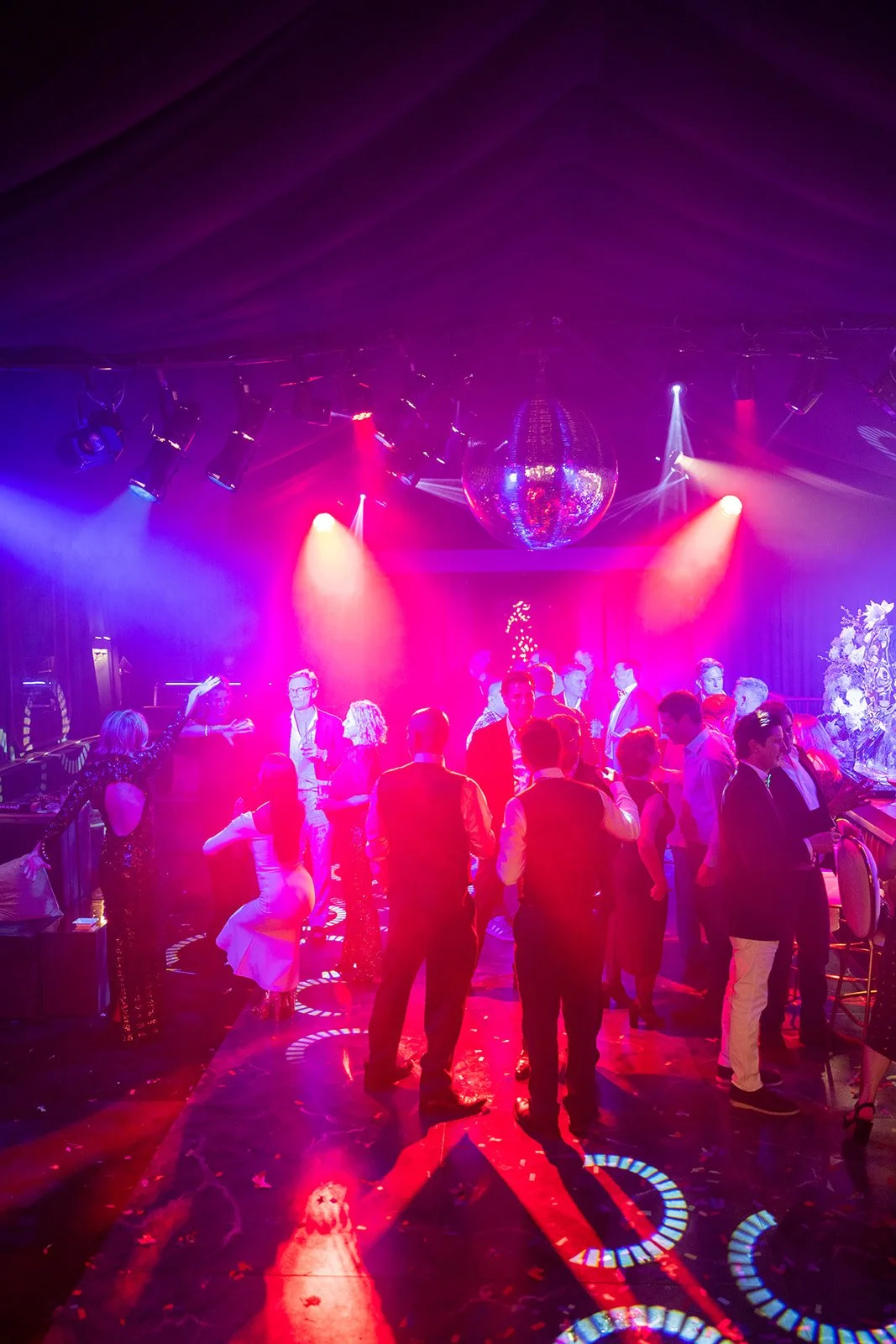 People dancing and socializing at a nightclub with colorful lighting and a disco ball.