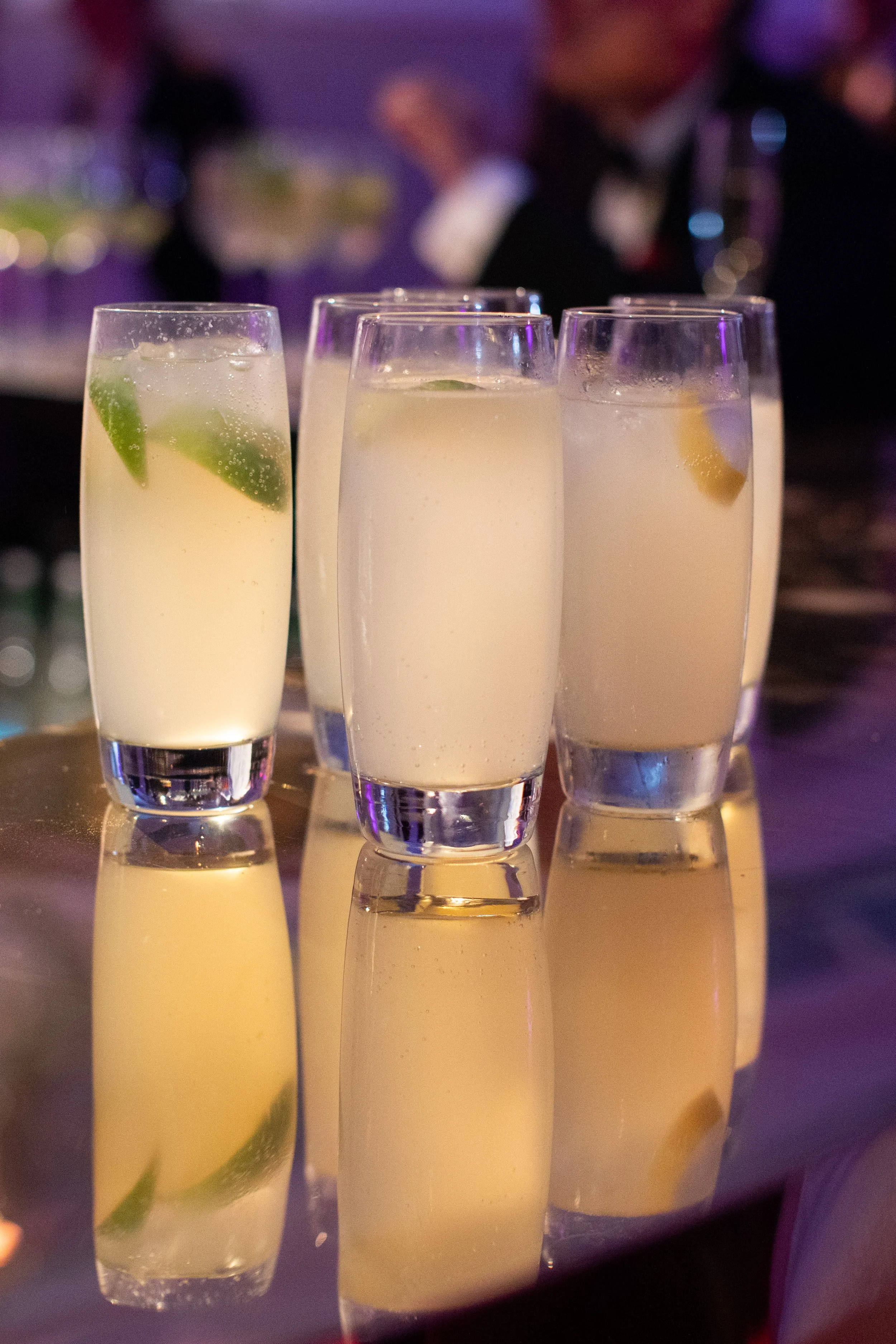 Four champagne flutes filled with a light yellow cocktail, garnished with lime or lemon wedges, on a reflective surface with a blurred background.