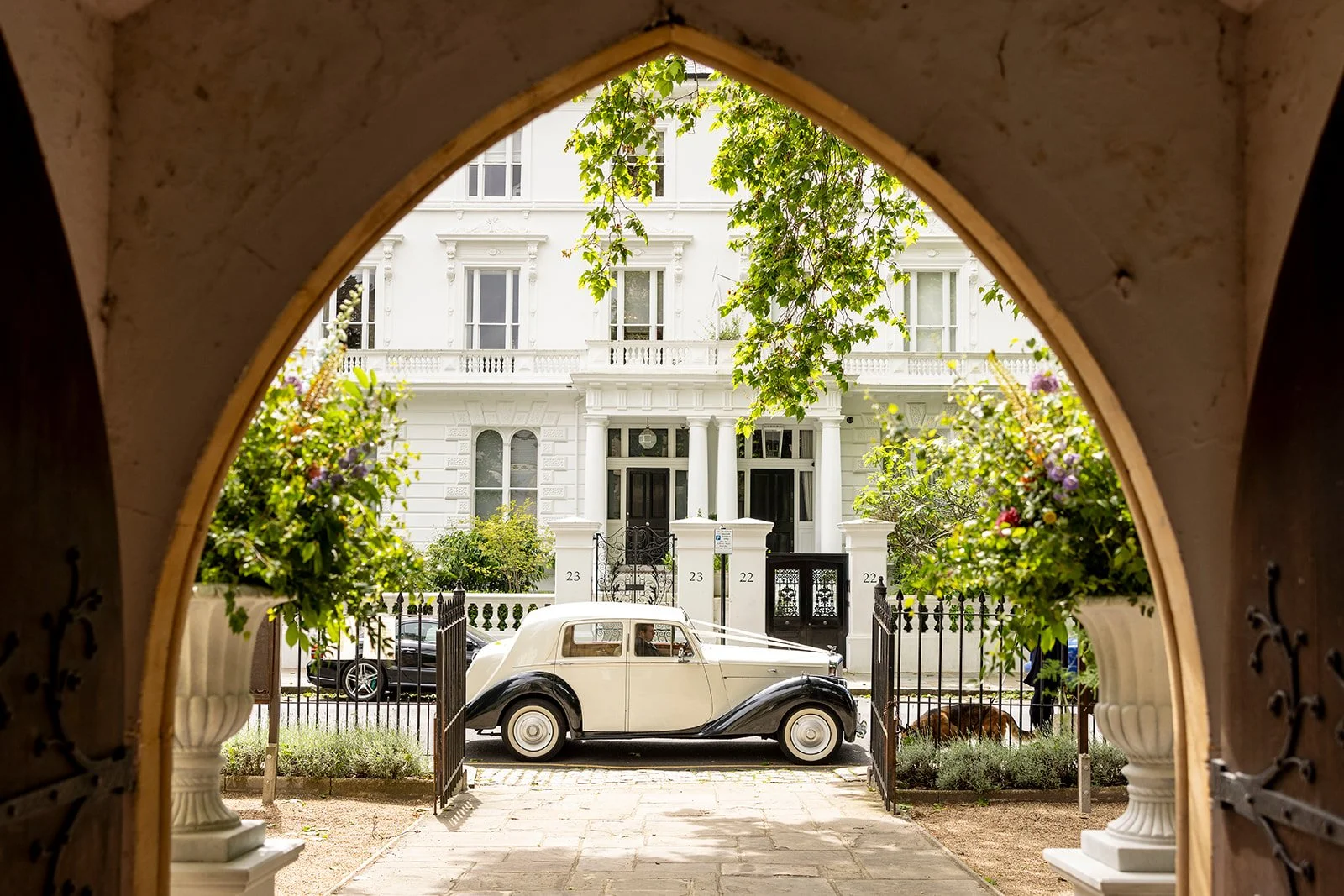 View of a vintage car parked in front of a white Victorian-style house through a decorative archway with flower pots on either side.