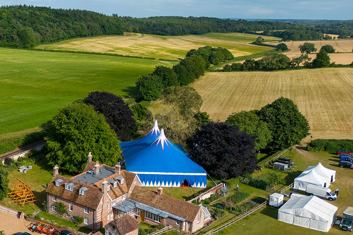 A large blue circus tent pitched in a rural area with traditional buildings, tents, and vehicles surrounding it. Green fields and trees stretch out into the distance.