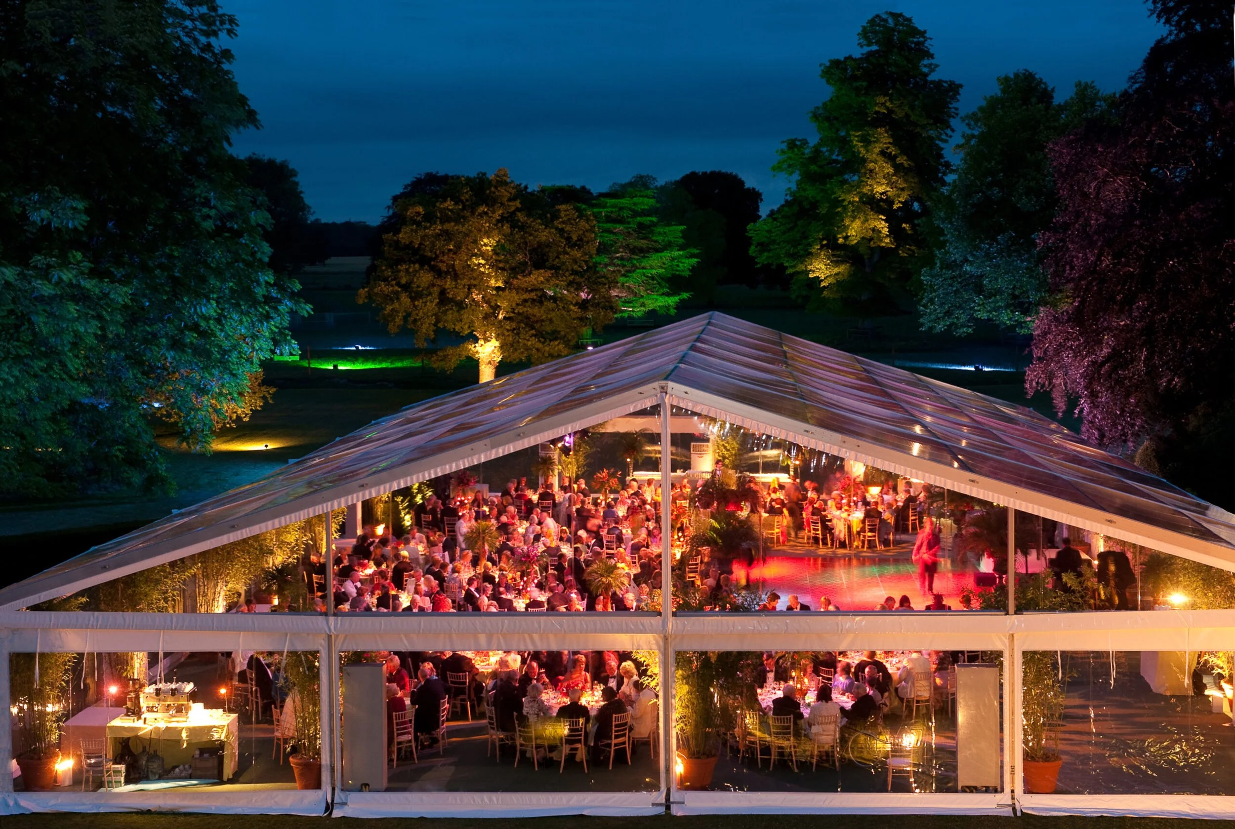 Nighttime outdoor event under a large transparent tent with guests seated at tables, illuminated by string lights, surrounded by lush green trees with colorful lighting.
