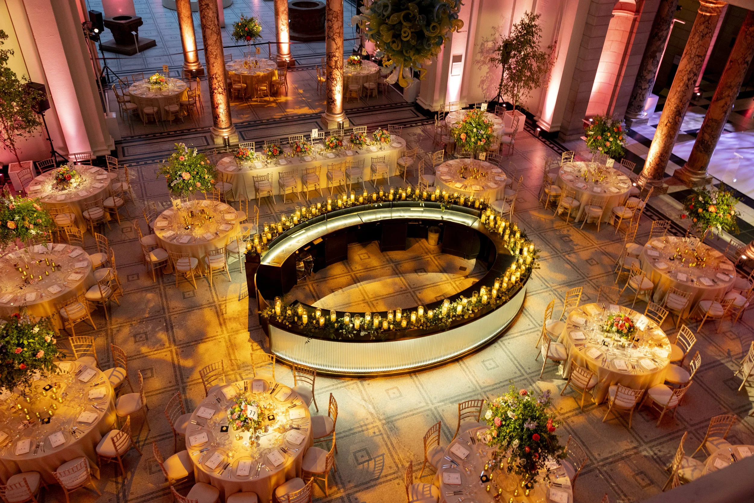 Elegant event space with round tables covered in pink tablecloths, decorated with floral centerpieces, gold chairs, and candles. There is a bar or DJ area with glowing lights surrounded by candles, large floral arrangements, and tall decorative colum