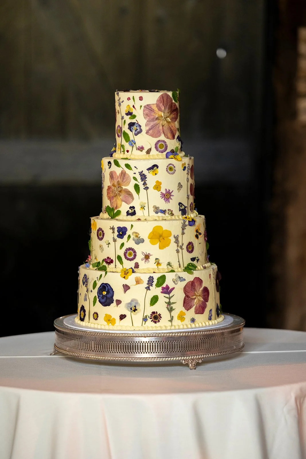 A four-tiered wedding cake decorated with edible flowers and greenery, placed on a silver cake stand on a round table with a white tablecloth.