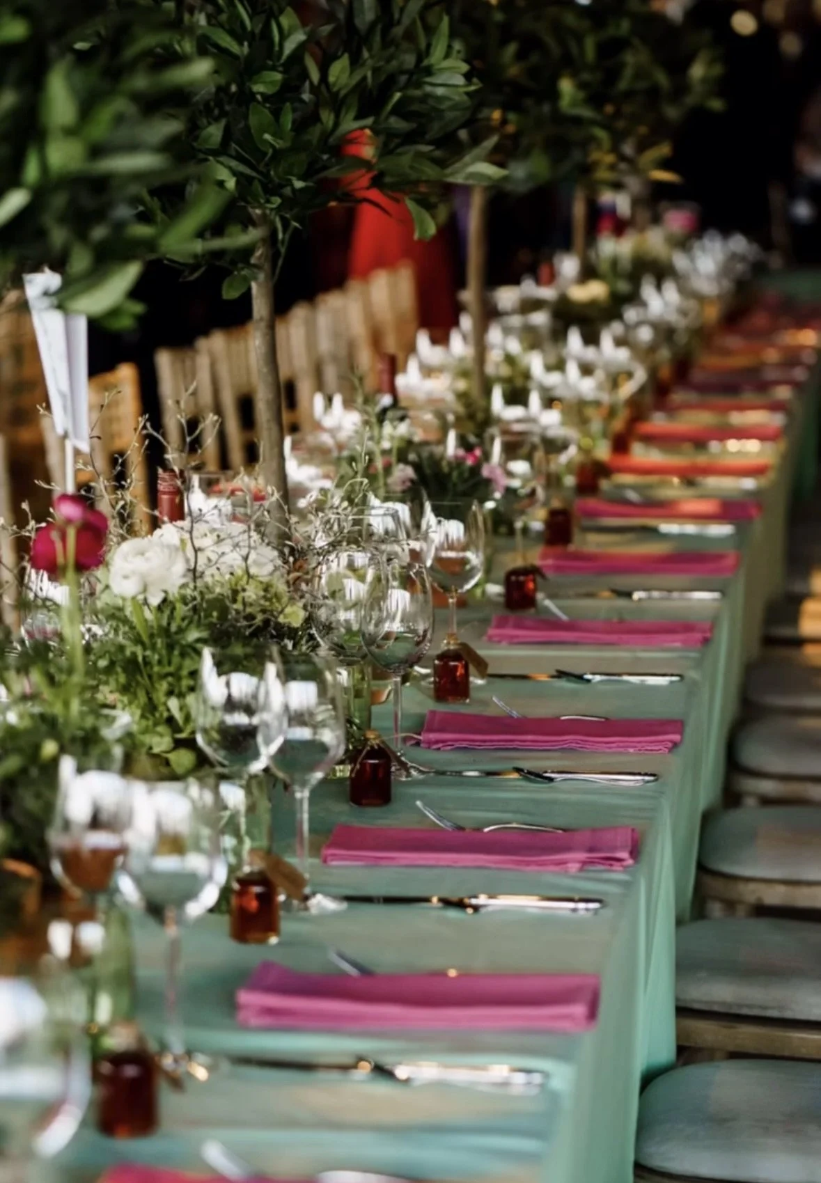 Elegant outdoor dining table set with pink napkins, glassware, and floral centerpieces, surrounded by greenery and chairs, for a special event or celebration.