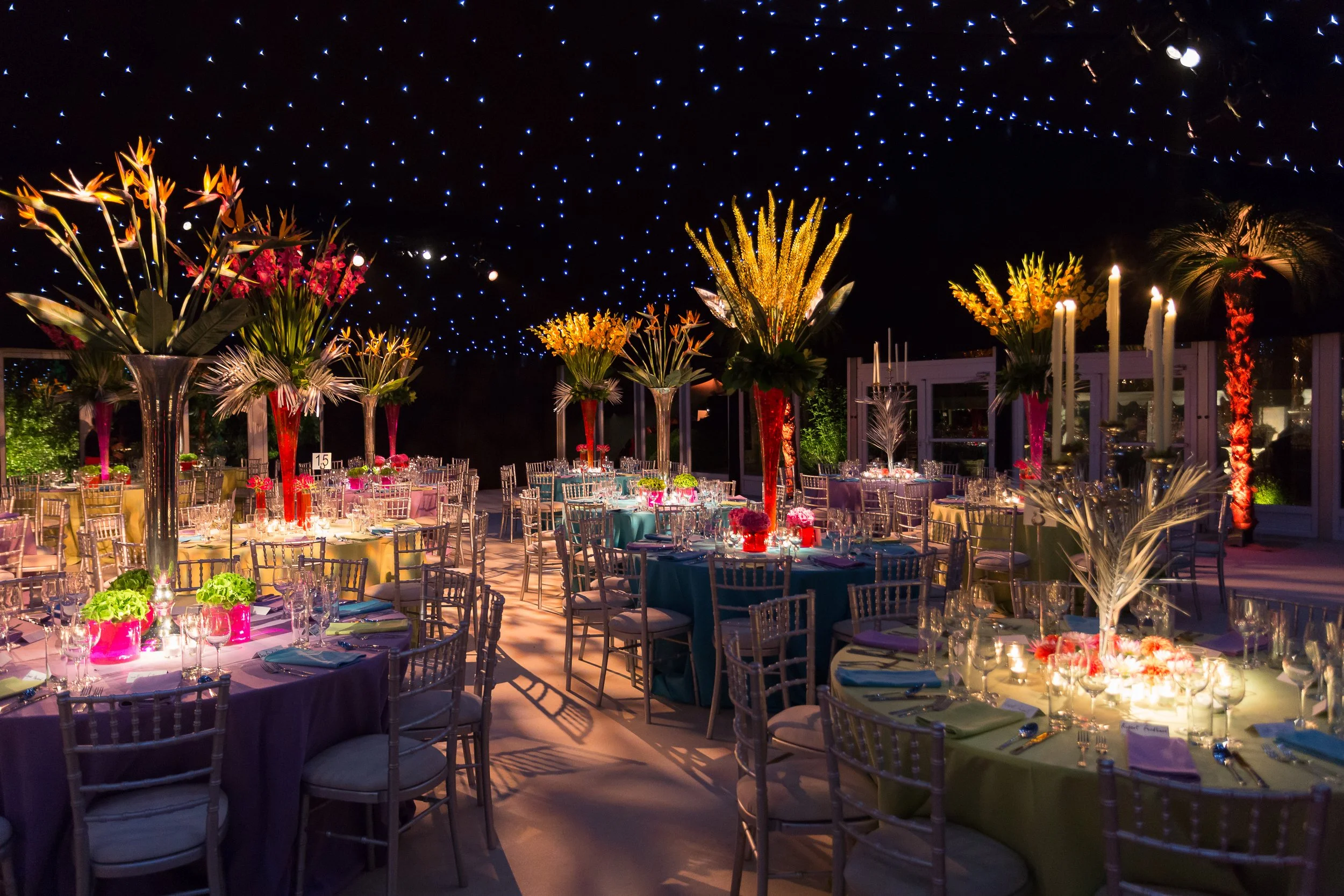 Elegant indoor event space decorated with tall floral centerpieces, candles, and string lights overhead, set up for a banquet or celebration.