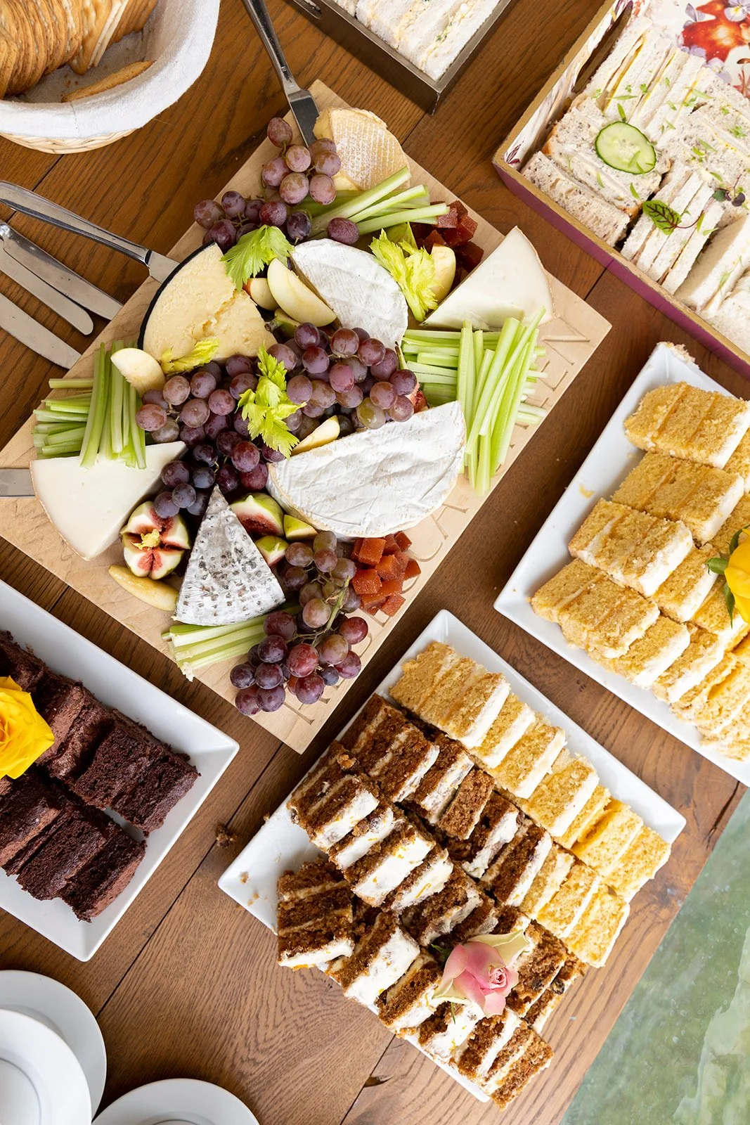 A cheese platter with grapes, apples, celery sticks, and various cheeses, surrounded by plates of sliced cakes and sandwiches on a wooden table.