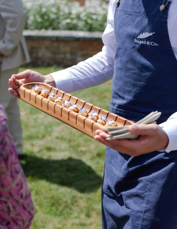 A person wearing a peaped & Co. apron holding a wooden tray with small appetizers or treats topped with blue and white toppings, outdoors on grass.