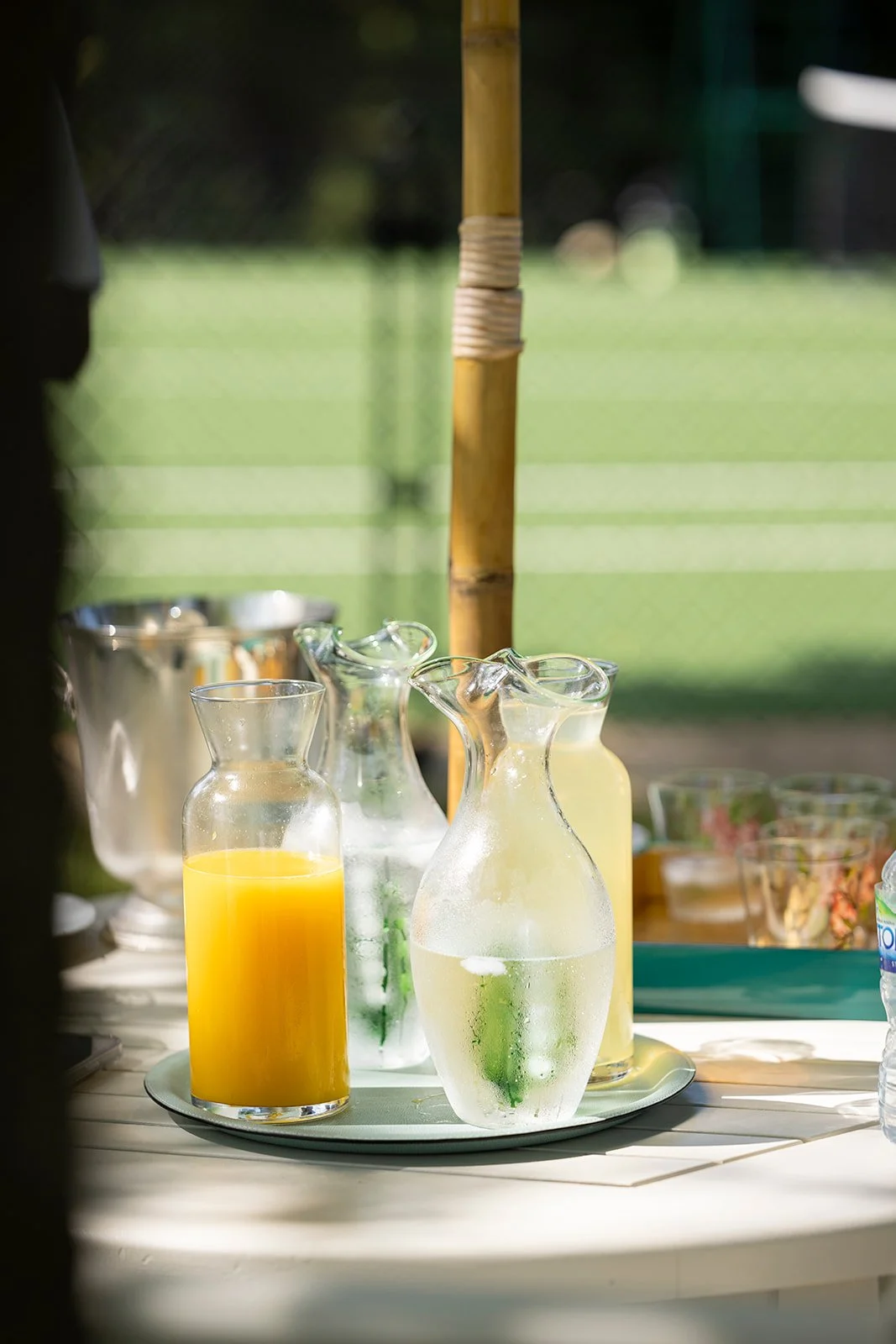 Glass pitchers containing orange juice and water, set on a tray outside, with a background of a sports field.