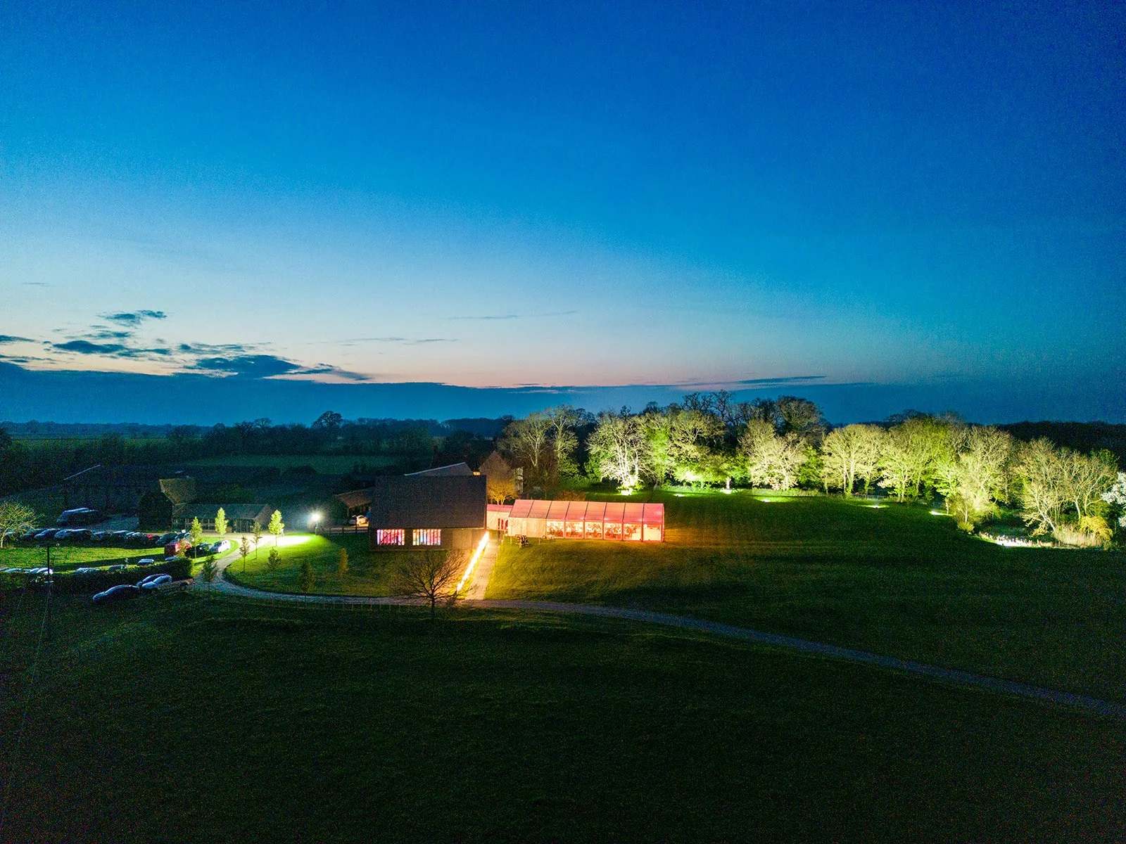 A farm landscape at dusk with a large grassy field, a barn, a greenhouse with red lighting, trees with illuminated trunks, and a parking lot with cars, under a sky turning dark blue.