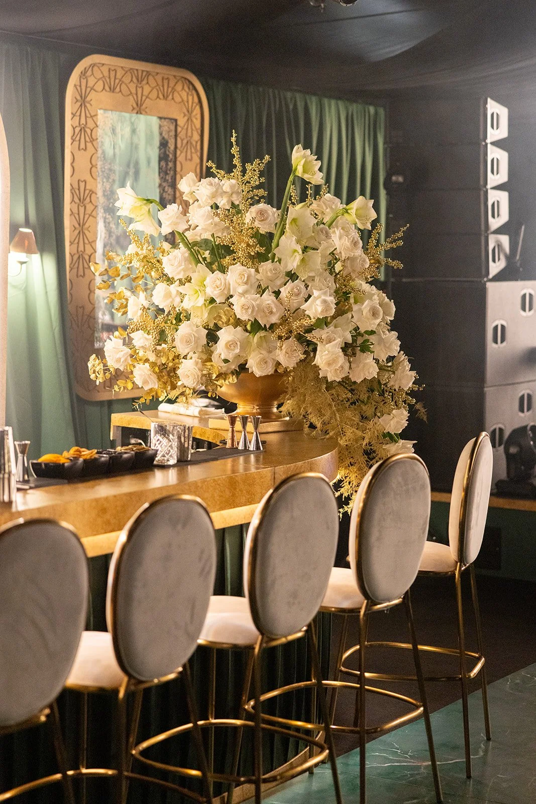 A large floral arrangement with white roses and lilies on a bar counter in a dimly lit venue with green curtains and sound equipment in the background.