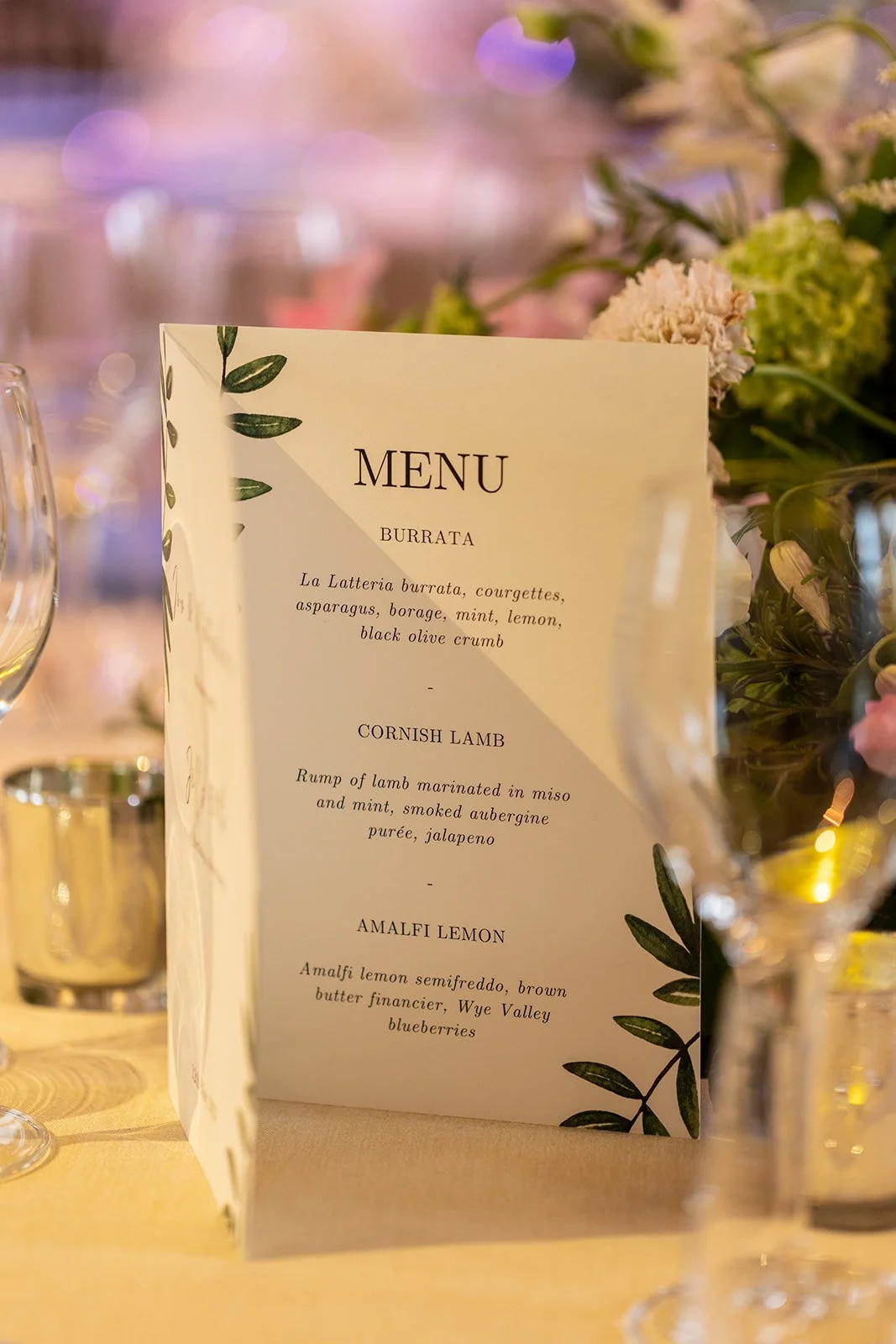 A printed menu with a decorative green leaf design, listing dishes including burrata, Cornish lamb, and Amalfi lemon, placed on a table with glassware and floral arrangements in the background.