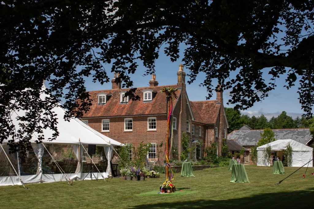 Outdoor wedding reception setup in front of a large brick house with tents, green tablecloths, and a decorated pole on a grassy lawn.