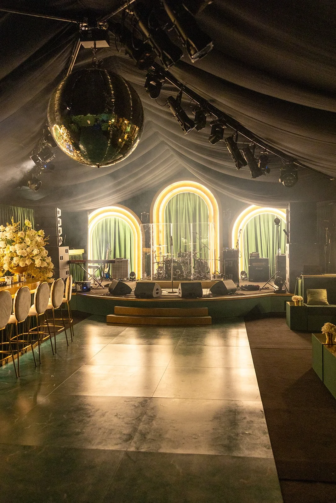 Empty stage with musical instruments and lighting, decorated with green curtains, neon arch accents, and surrounded by chairs and floral arrangements in a dimly lit venue with a disco ball hanging from the ceiling.