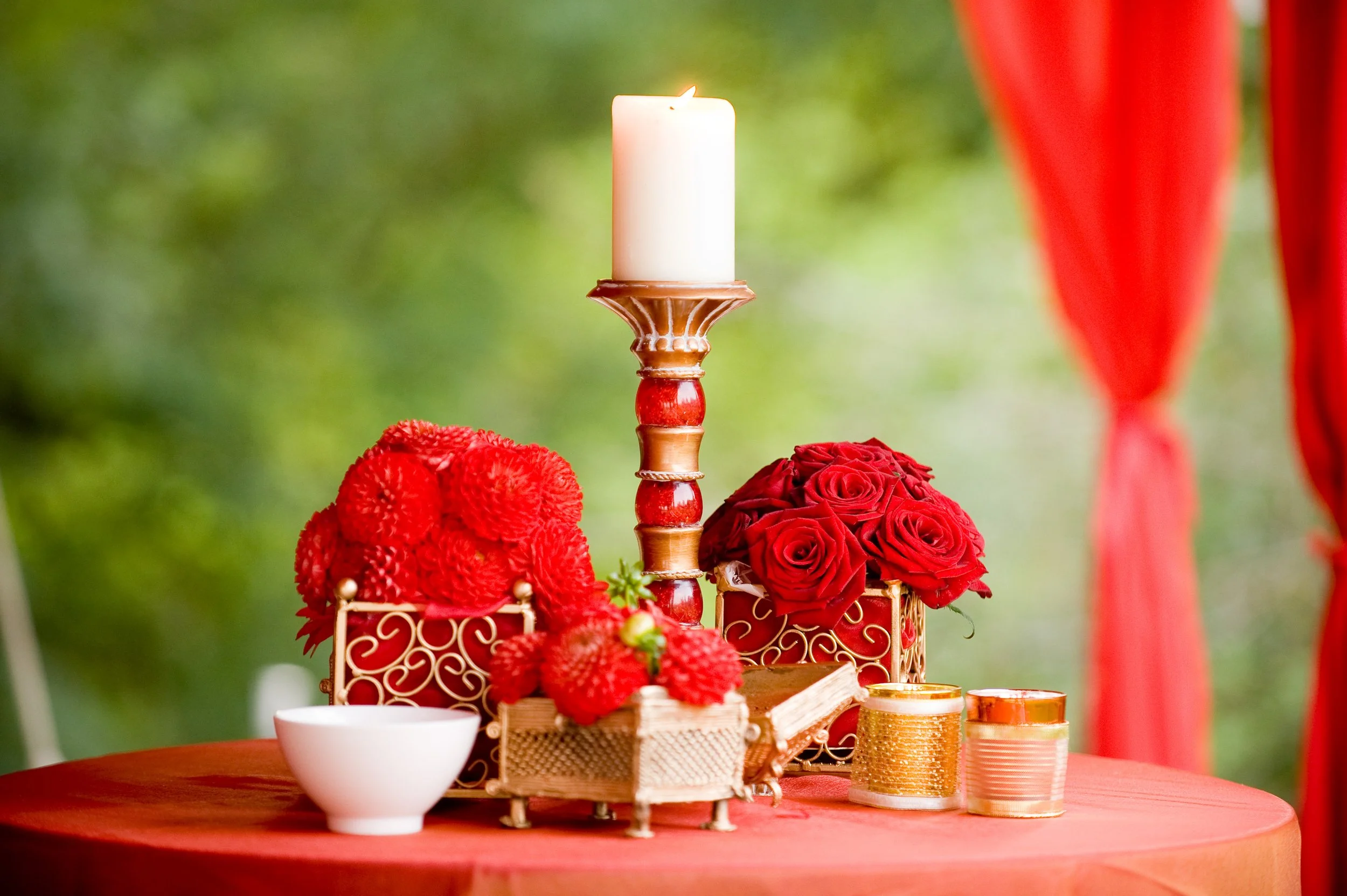 A decorative candle on an ornate stand surrounded by red flowers and gold accents on a red tablecloth, with a blurred outdoor background.