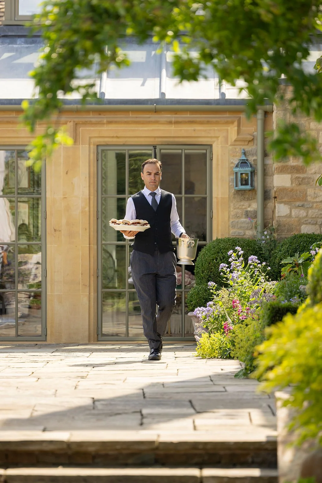 A man in formal attire, carrying a tray of sandwiches and a wine bucket, walking outside a house with large windows and a garden.