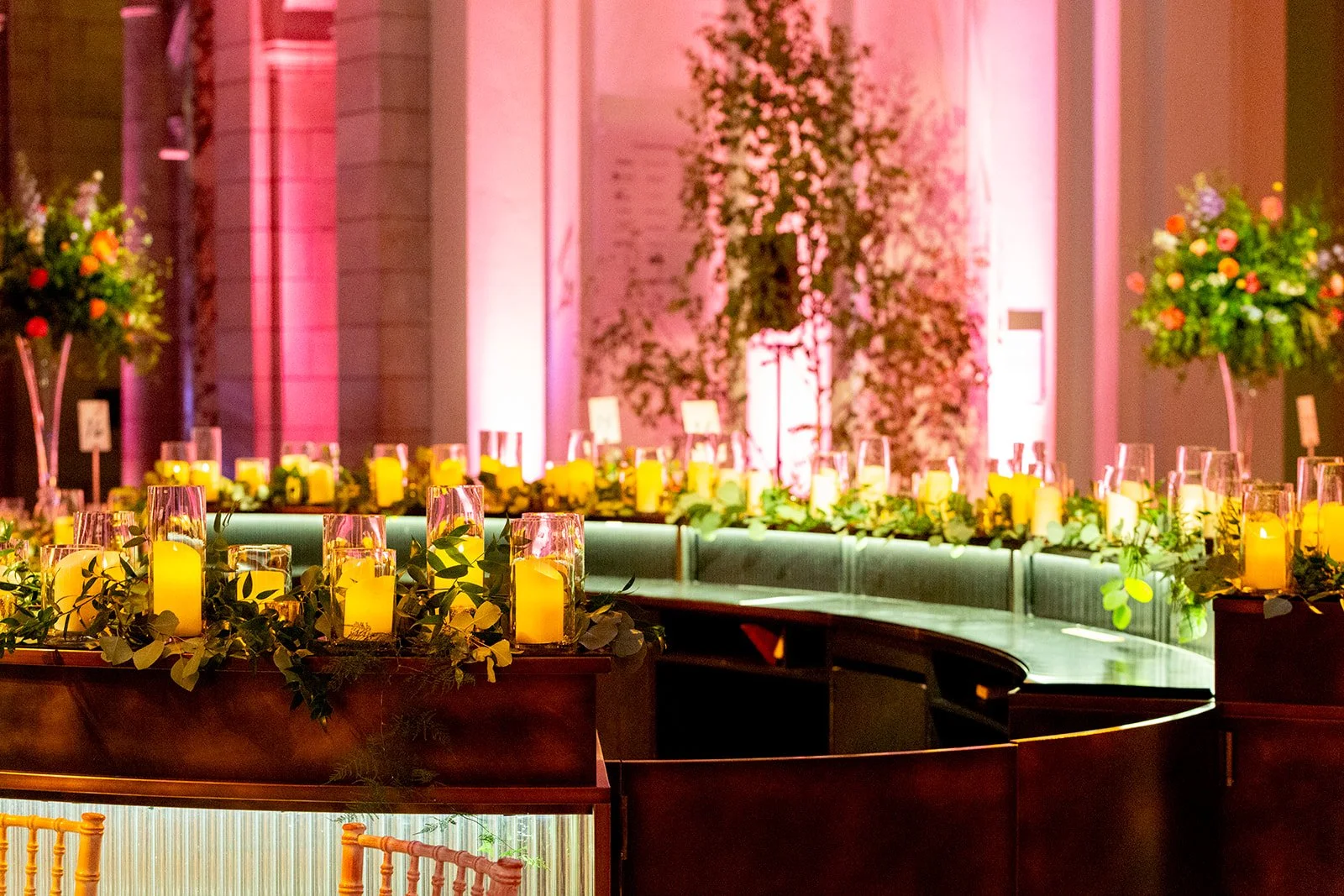 Decorated banquet table with yellow candles, greenery, and floral arrangements in a dimly lit, elegant event space with pink and purple lighting.
