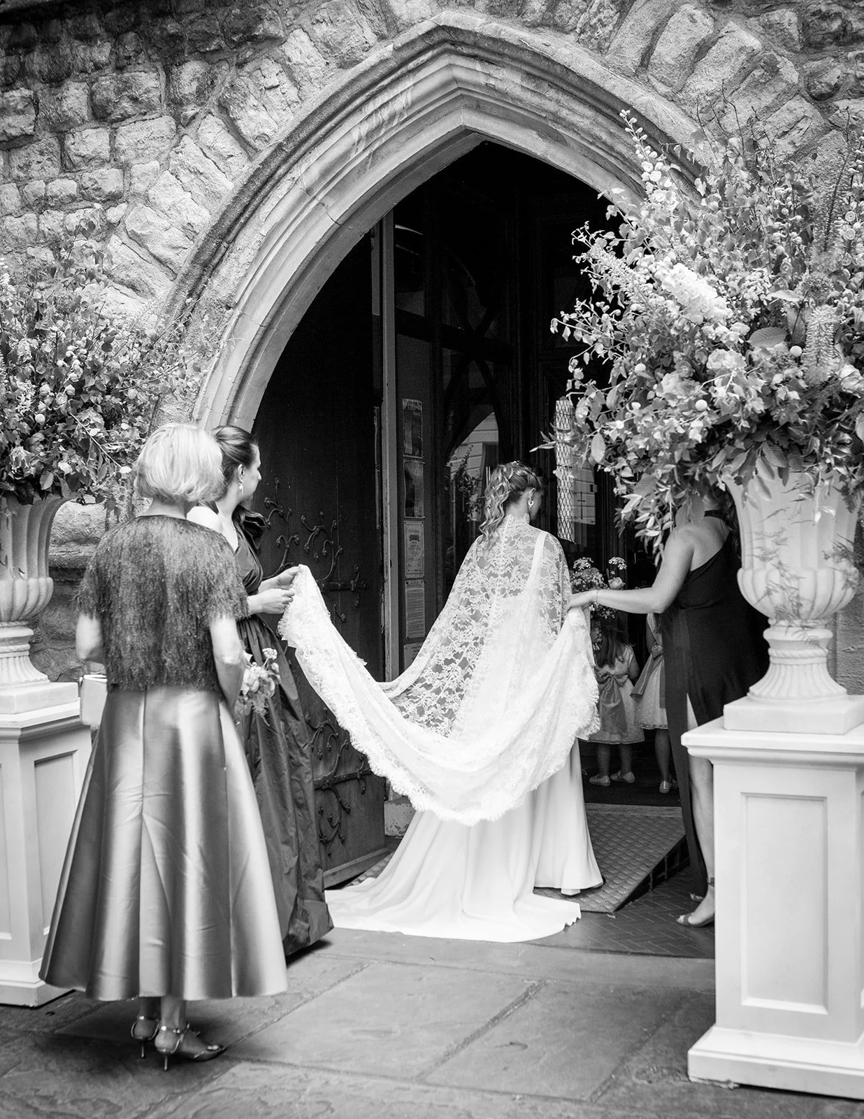 Black and white photo of a bride with a long lace train entering a church, with three women assisting her, two holding her train and one at her side, near large floral arrangements outside the church doorway.