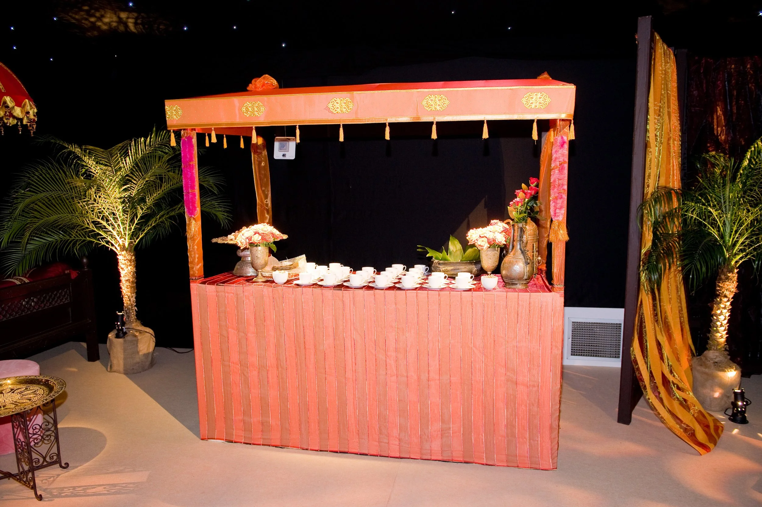 Decorative outdoor setting featuring a pink striped bar with teacups, flower arrangements, and plants on either side, with a starry night sky in the background.