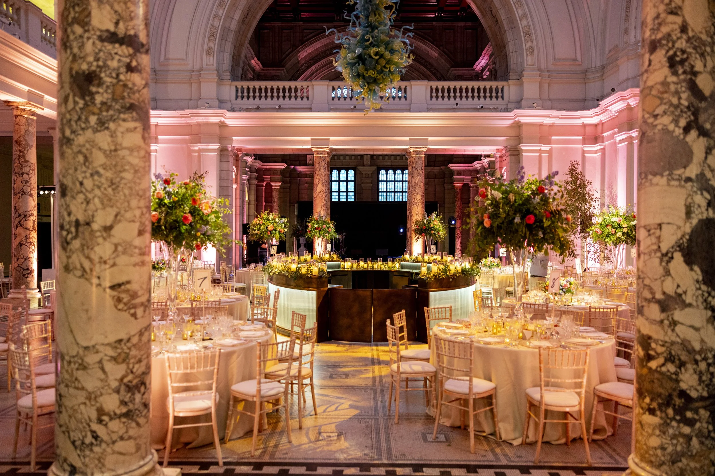 Elegant banquet hall decorated with pink lighting, marble columns, flowers, and a chandelier in the center.