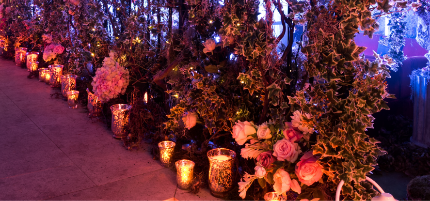 Decorative outdoor walkway with lanterns, flowers, and greenery illuminated by colorful lighting.