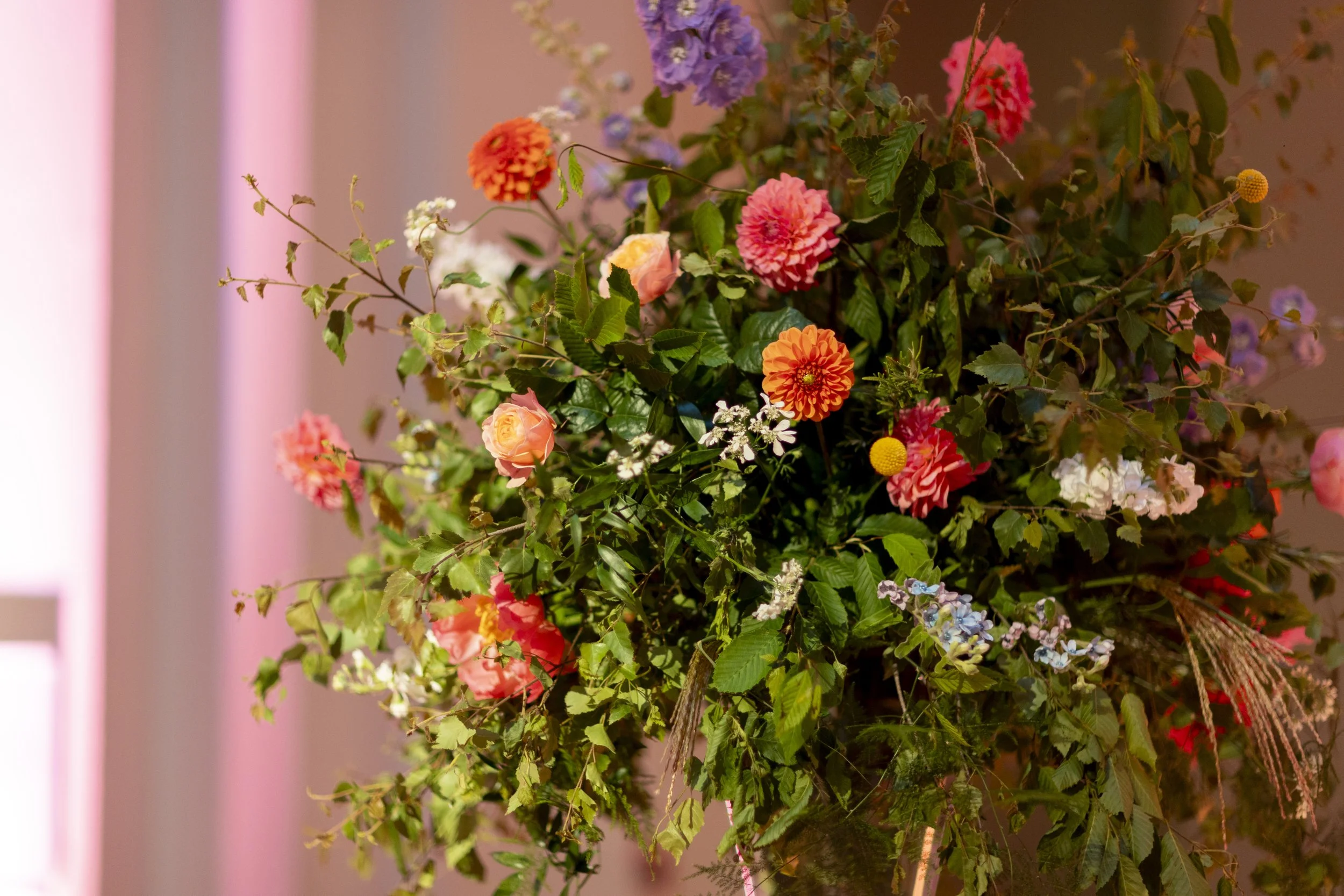 A large arrangement of colorful flowers including pink, orange, purple, yellow, and white blossoms with green leaves, set against a blurred background.