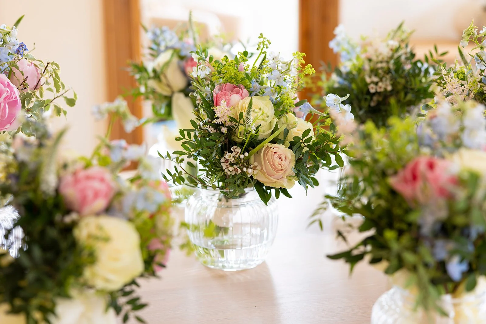 Floral arrangement with pink, white, and light blue flowers in a clear glass vase on a wooden table, with more arrangements blurred in the background.