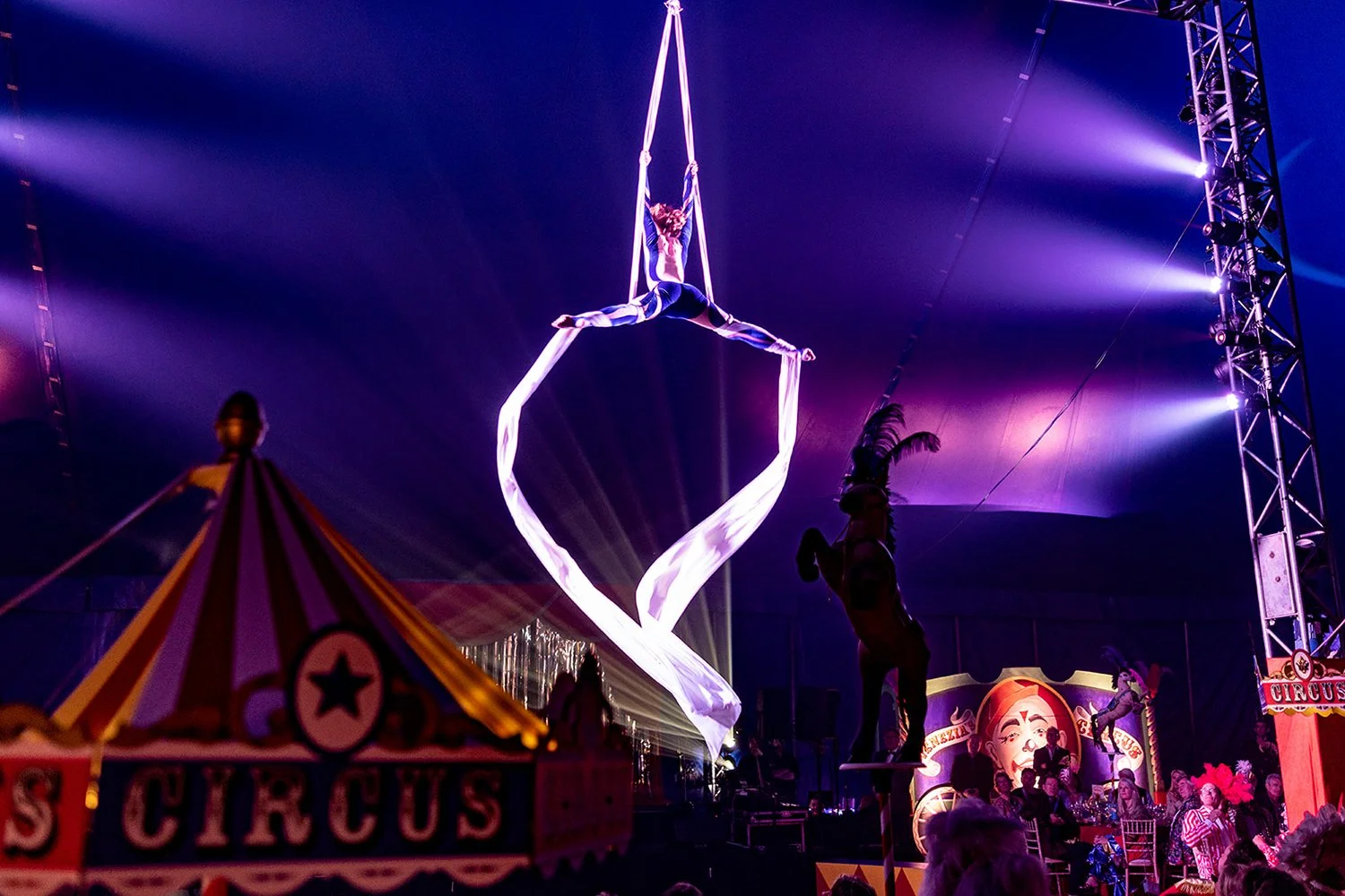Aerial silk performers in a circus tent, with one performer hanging upside down and another standing on a platform, illuminated by purple and blue stage lights, circus signs, and an audience seated at tables.