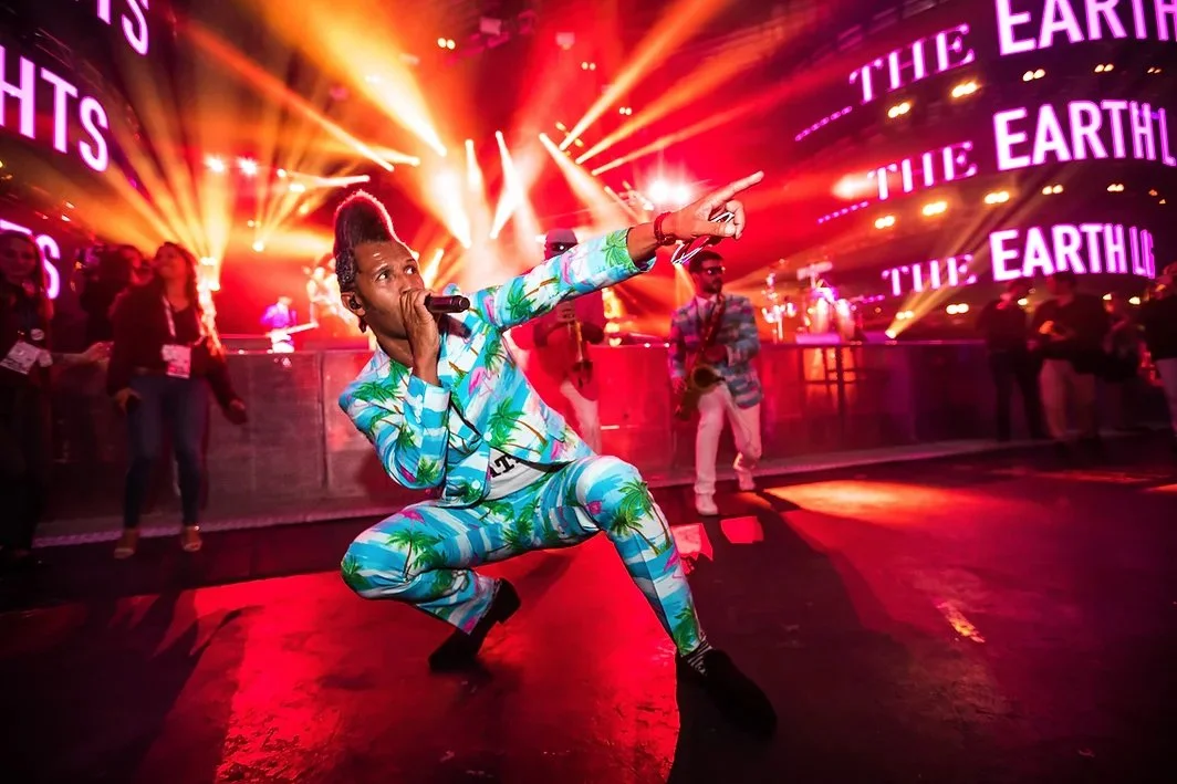 Performer in a colorful, tropical print suit kneeling on stage with a microphone, pointing outward, during a concert with vibrant stage lights and people in the background.