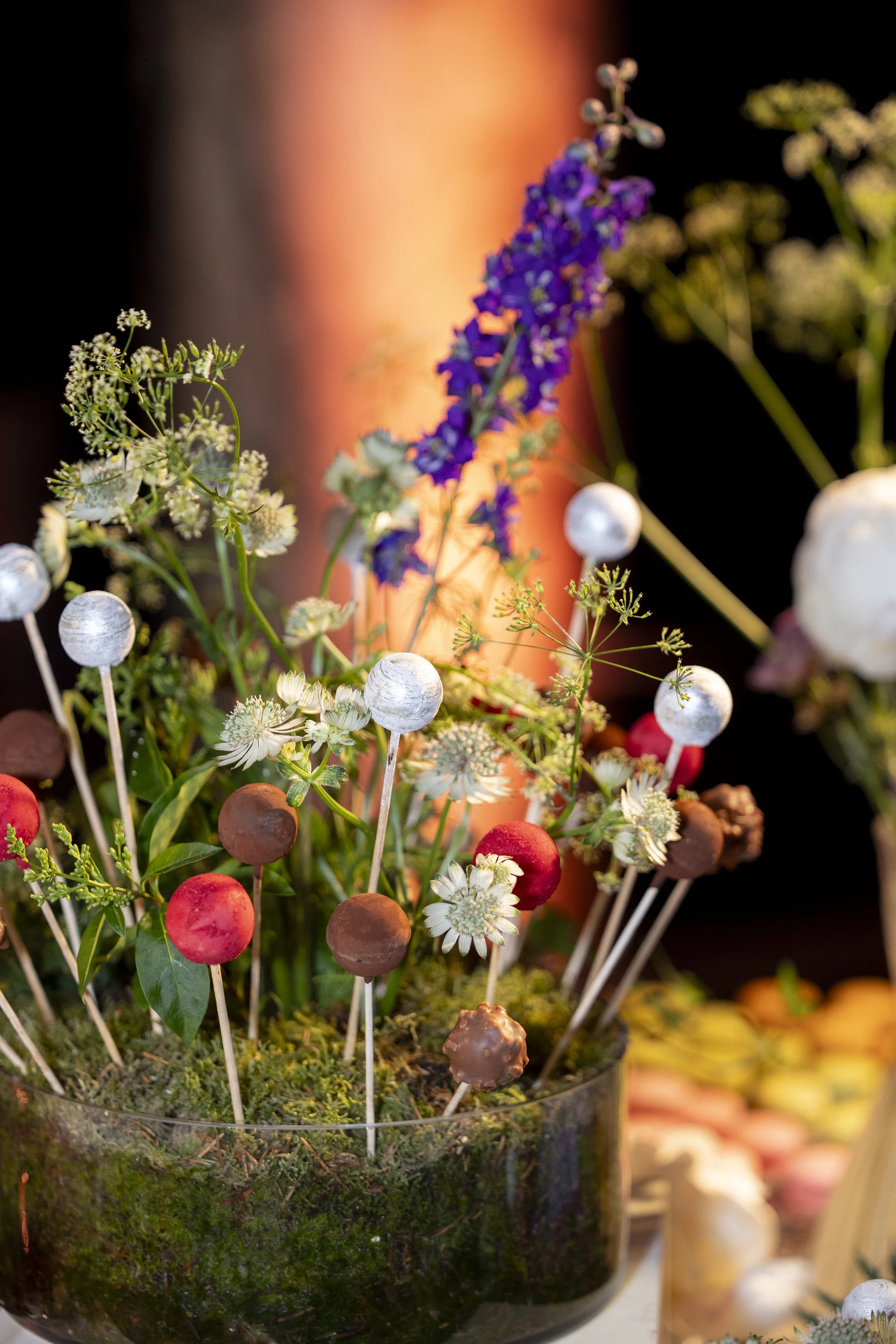 Decorative arrangement of small white, purple, and red spherical ornaments on sticks with greenery and white flowers in a glass container.