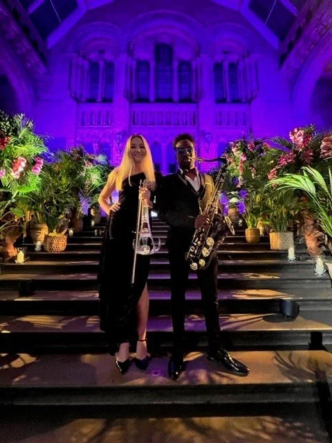 Two musicians, a woman and a man, stand on stairs in front of a decorated stage with lots of greenery and flowers, in a grand cathedral-like venue illuminated with purple lights.