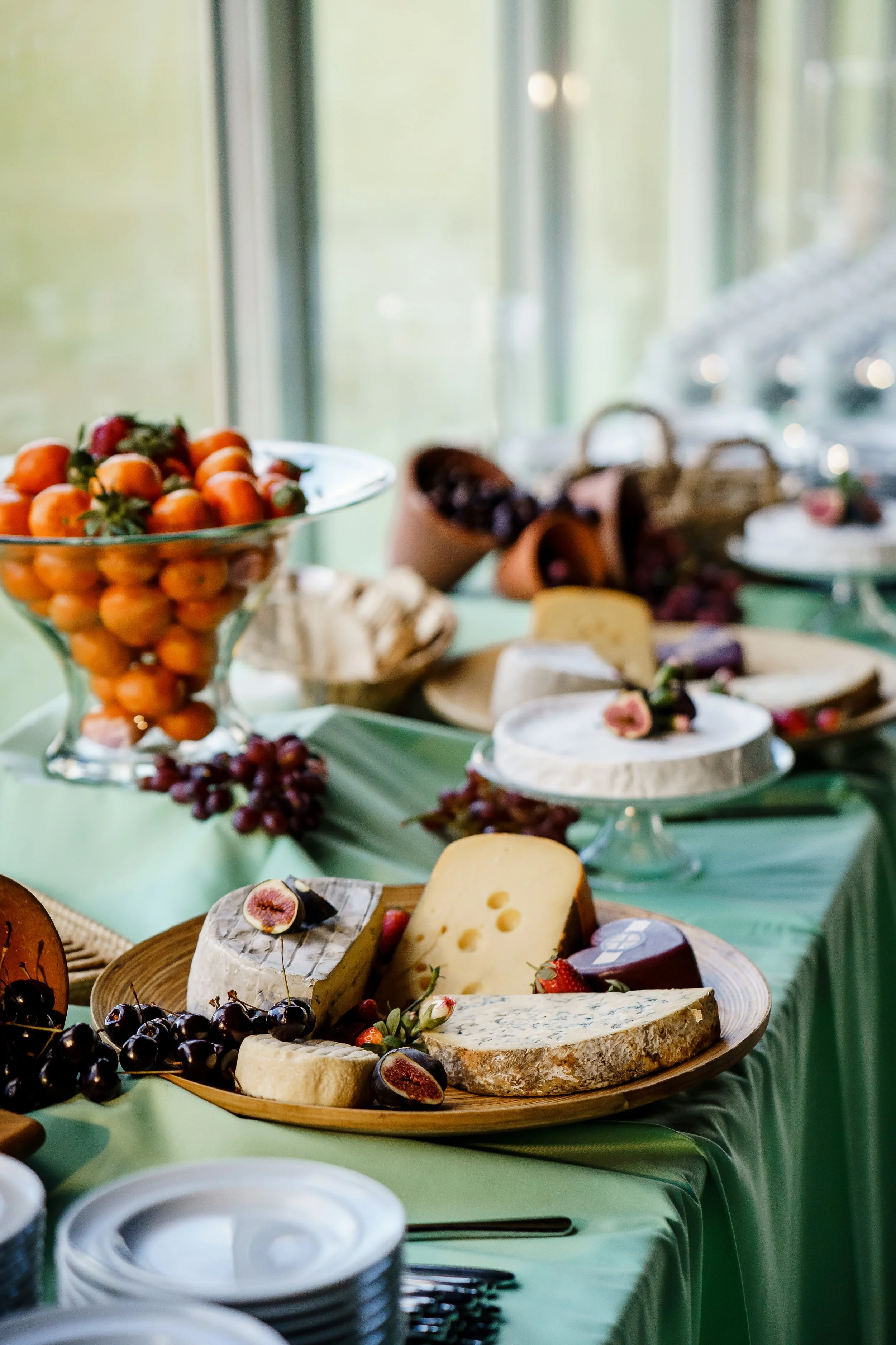 A display of various cheeses, fruits, and desserts on a table with a green tablecloth, set near a window with natural light.