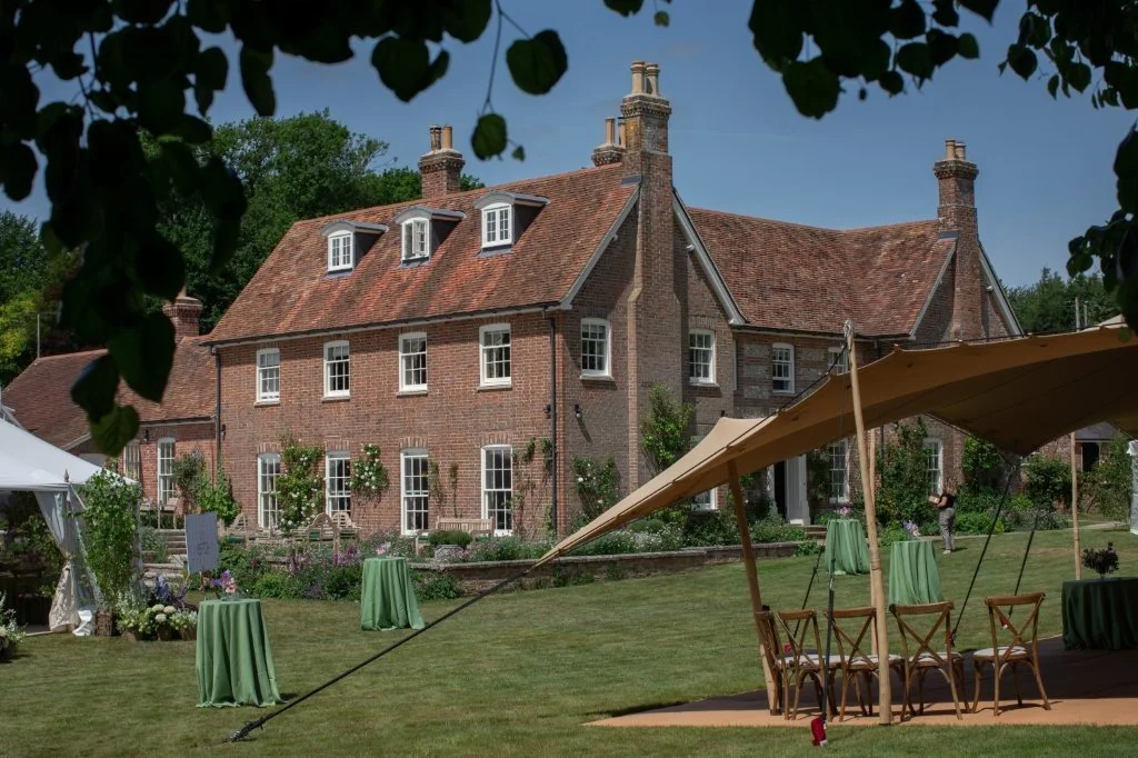 Large brick house with a red tile roof, multiple chimneys, and several dormer windows in a garden setting. Tents and tables with green tablecloths are set up on the lawn, likely for an outdoor event, with trees and a clear blue sky in the background.