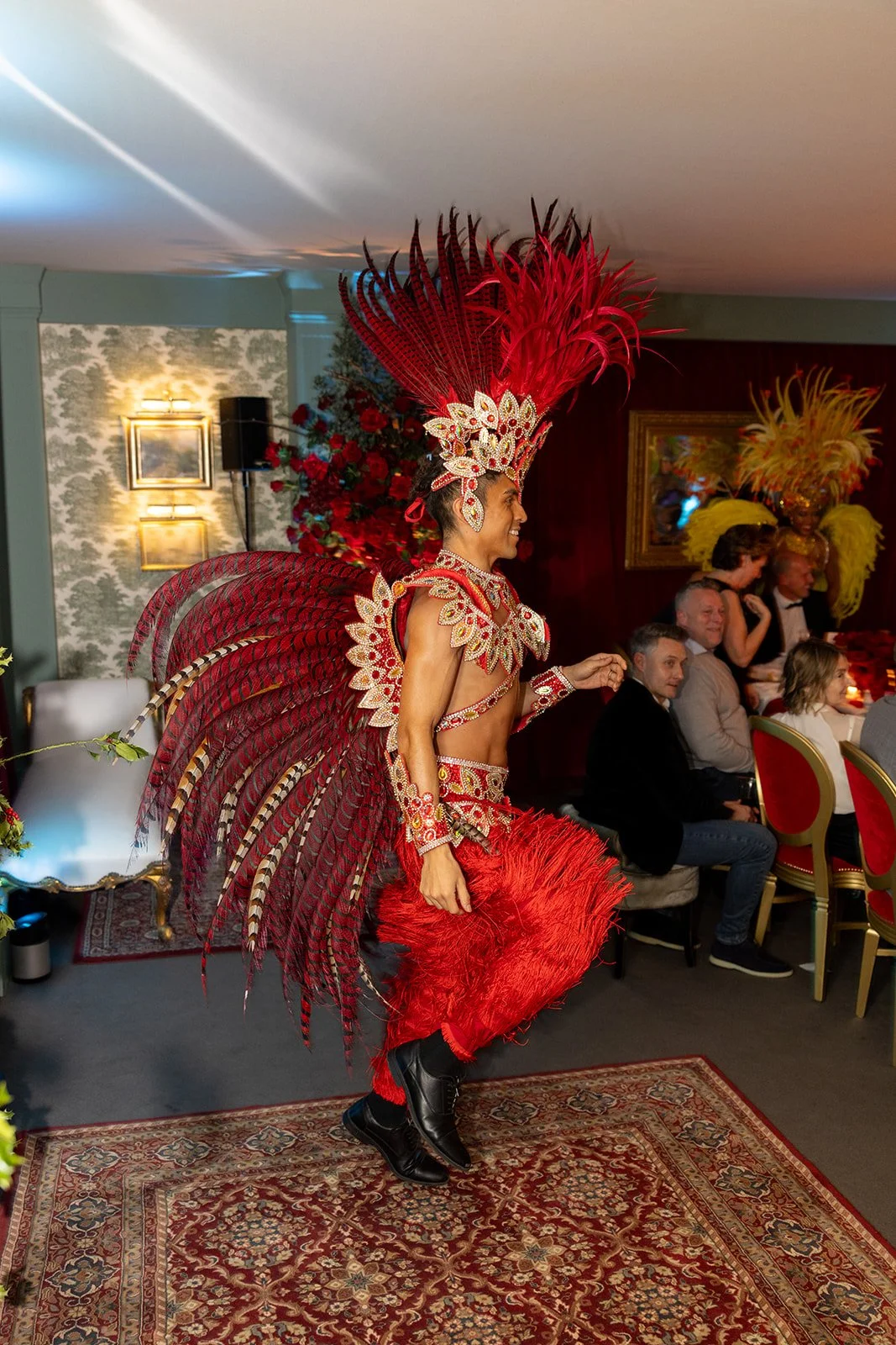 Performer in elaborate red and gold costume with feathers and sequins, holding a red feathered prop, in a decorated indoor setting with seated audience.