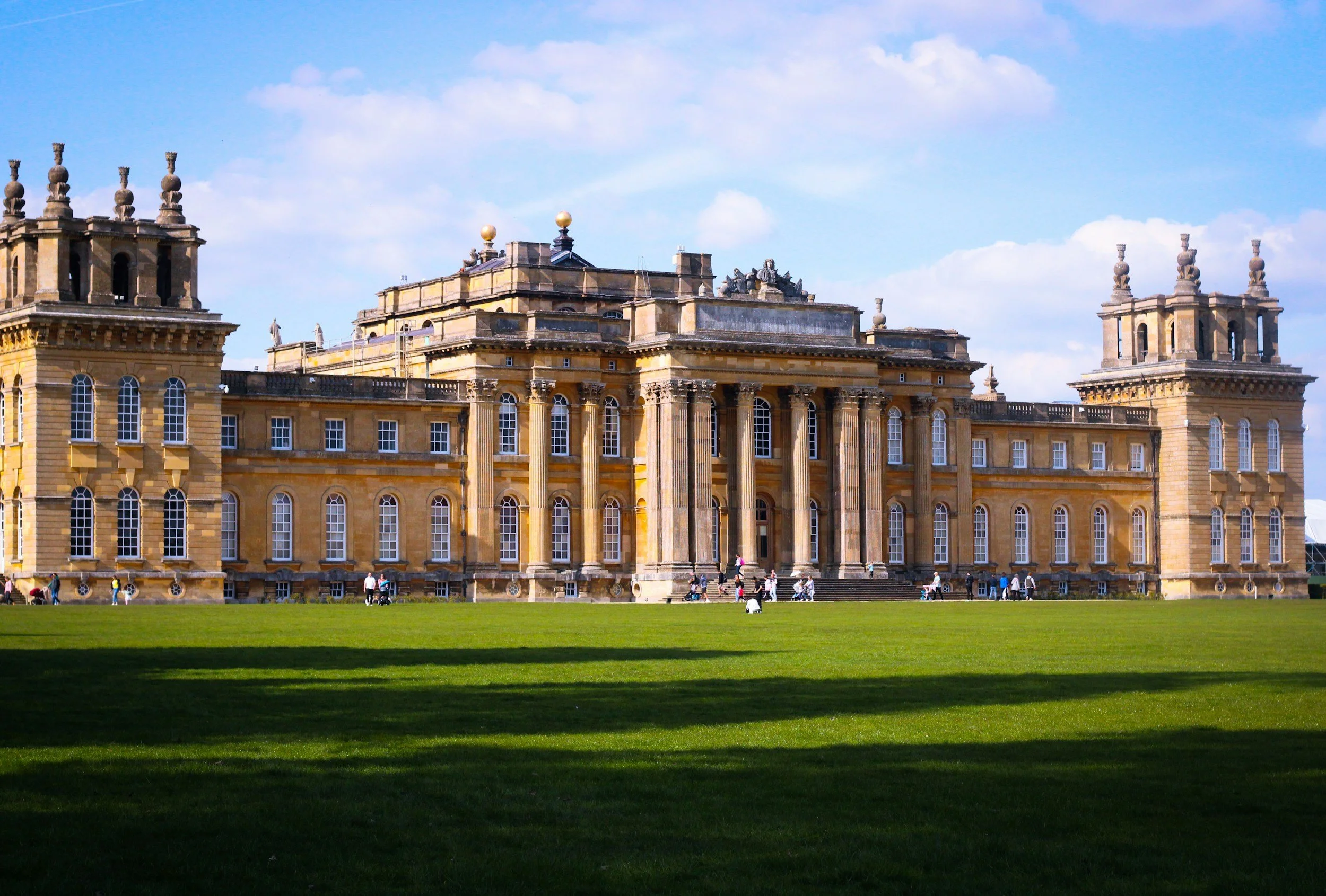 A large historic castle with a yellowish stone facade, multiple windows, and ornate architectural details. People are walking on the lawn in front, under a partly cloudy sky.