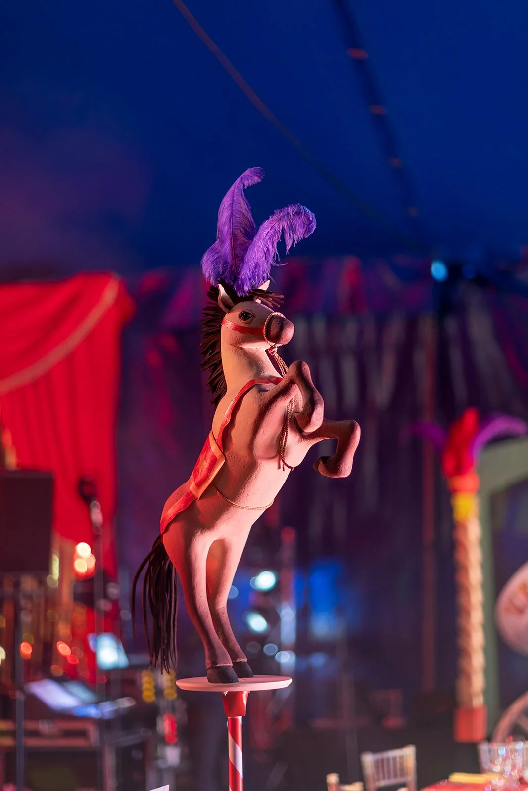 A toy carousel horse with a pink body, black mane, and purple feathers on its head, set in a colorful circus tent environment.