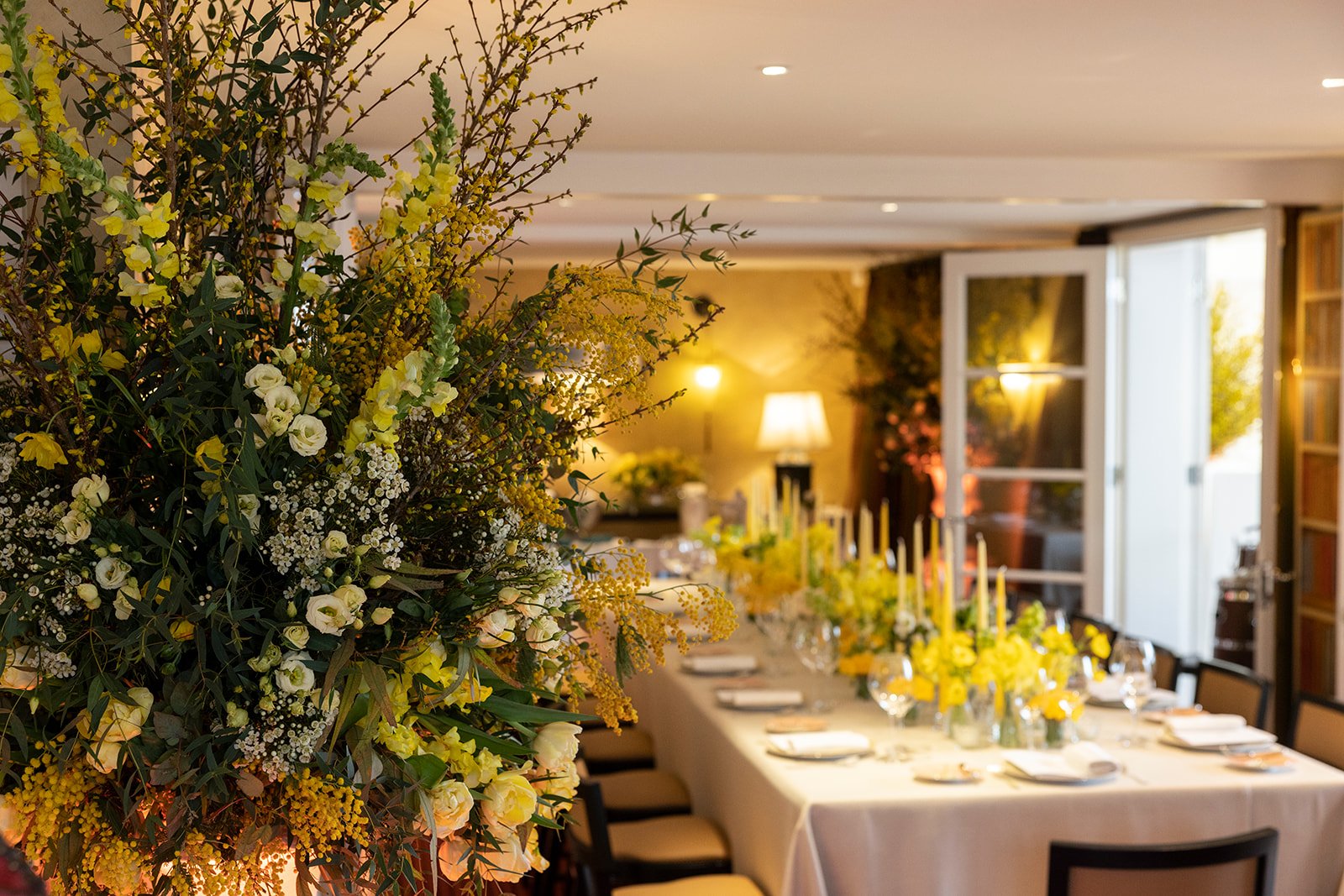 A long dining table decorated with yellow flowers and candles in a warmly lit room, with an open door leading to an outdoor area.