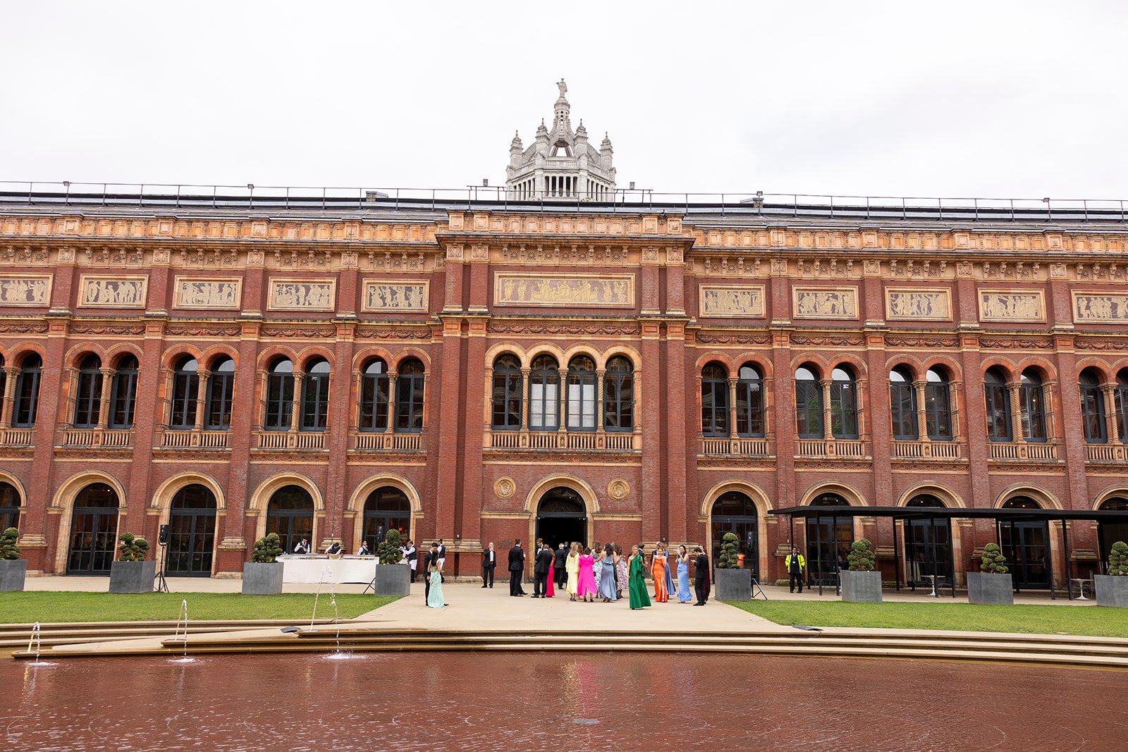 People dressed in formal attire gather outside a grand old red brick building with arched windows and decorative gold accents, with a fountain in the foreground and a cloudy sky overhead.