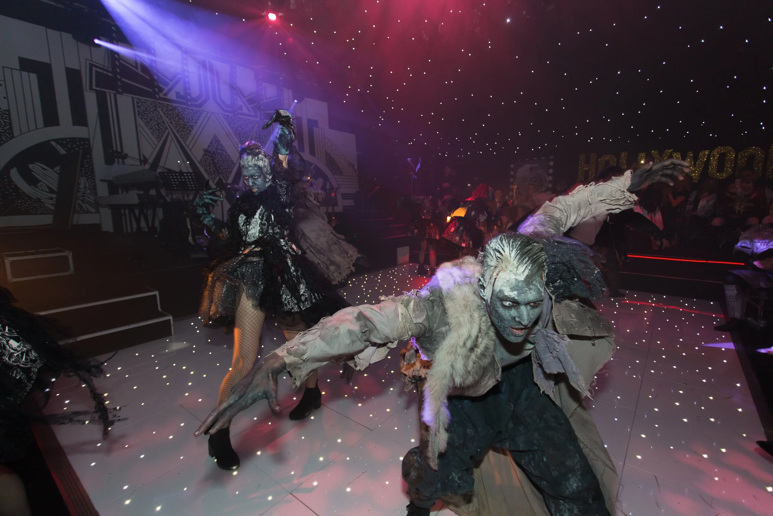 Performers on stage dressed as monsters and creatures with horror-themed makeup, costumes, and sculptures, under starry ceiling lights at a Halloween event or costume party.