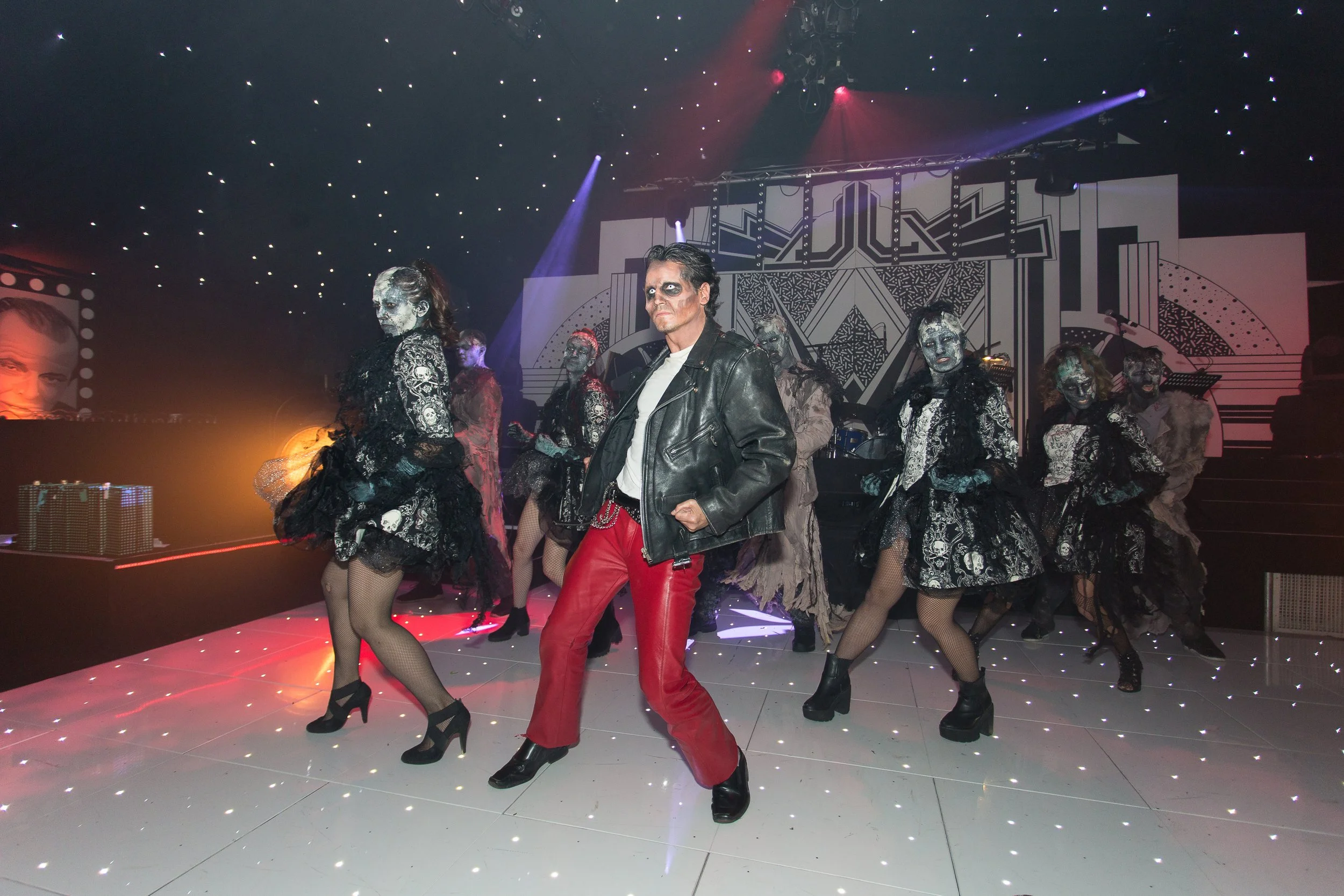 A performer in a black leather jacket and red pants standing center stage, surrounded by dancers in black laced dresses with skull patterns, wearing fishnet stockings and high heels, in a performance setting with stage lights and geometric backdrop.