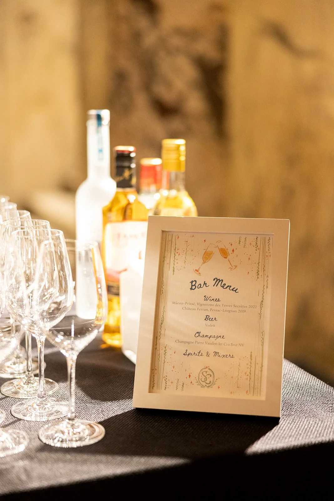 A table setup with empty wine glasses, bottles of alcohol, and a framed menu listing wines, beer, champagne, and spirits at an event or celebration.