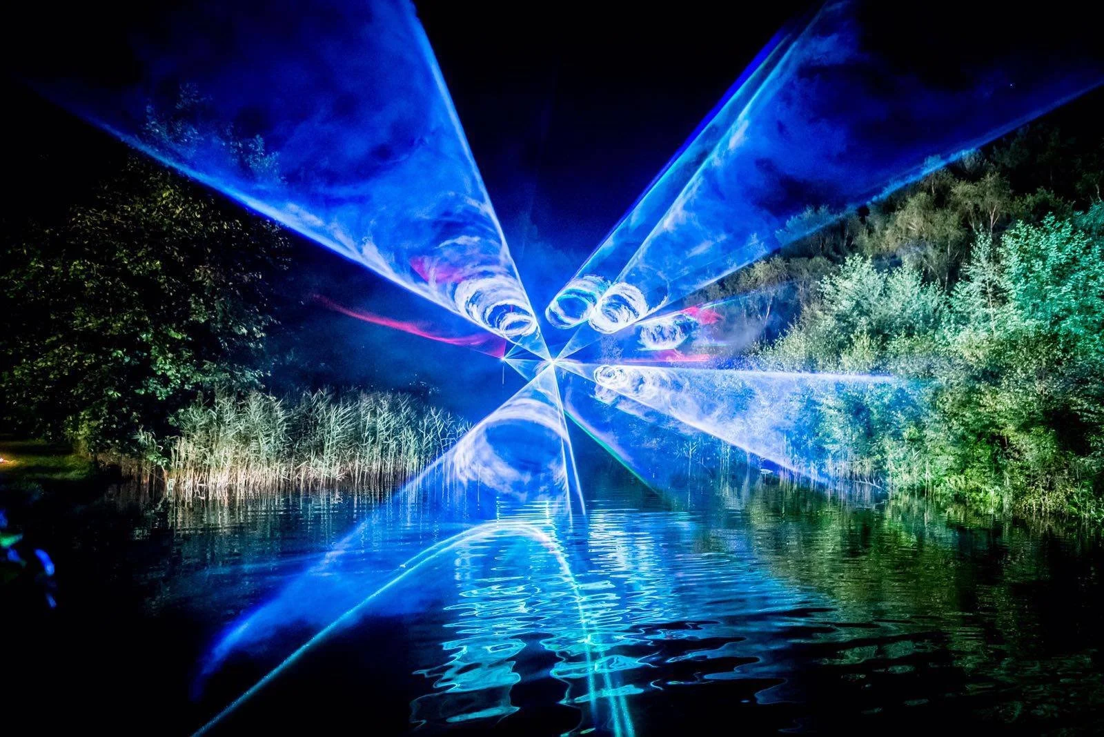 Blue laser light show over a lake at night, with reflections on the water surface and trees on both sides.