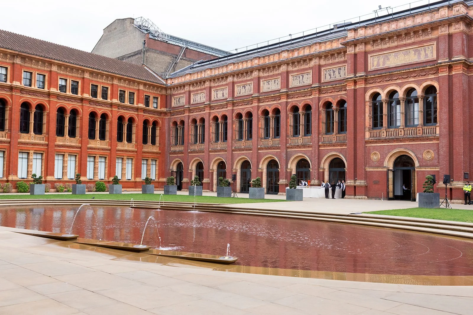 Red brick building with arched windows, a fountain in the foreground, and some people near the entrance.
