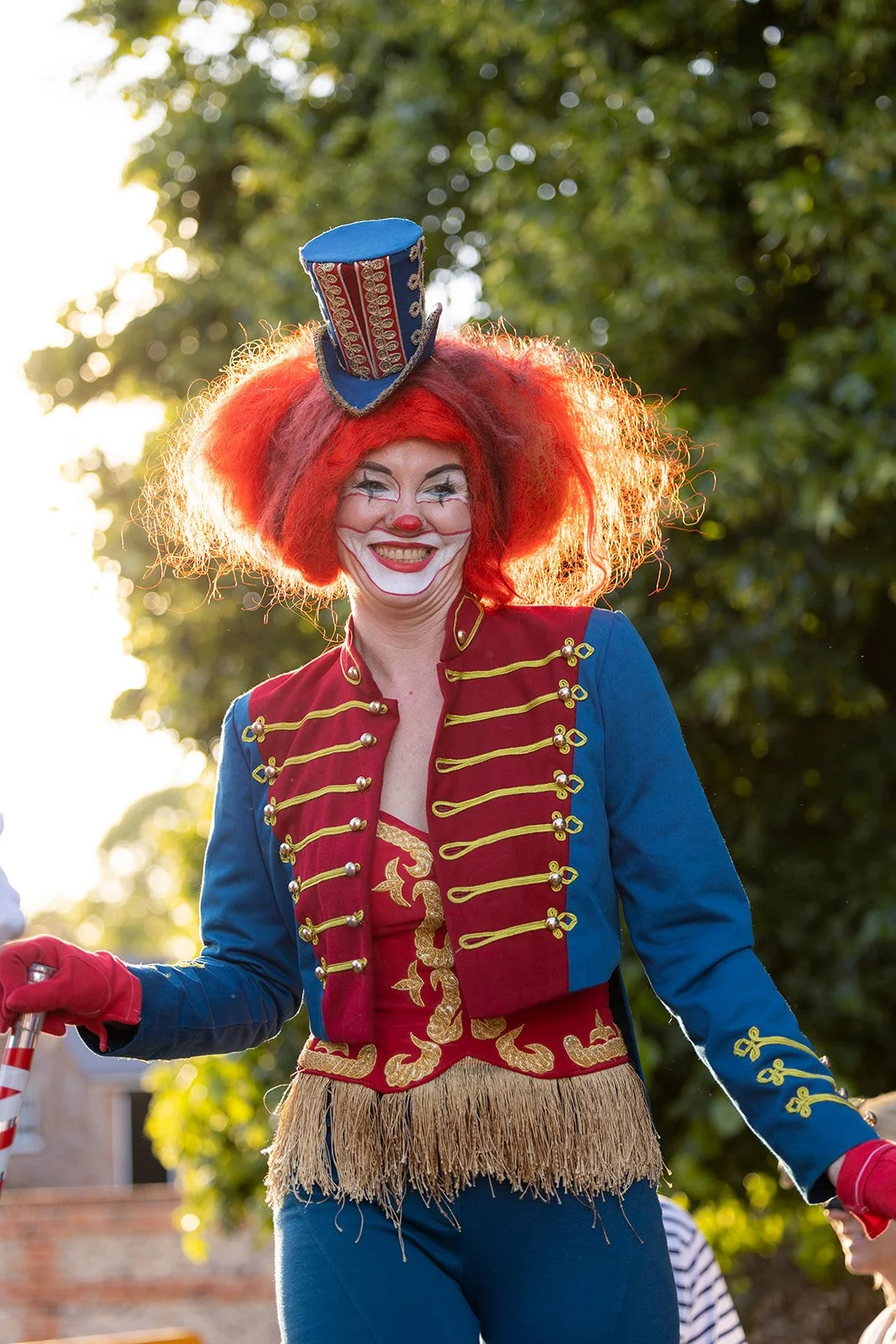 A woman dressed as a circus clown with red hair, face paint, and a colorful costume, smiling outdoors.