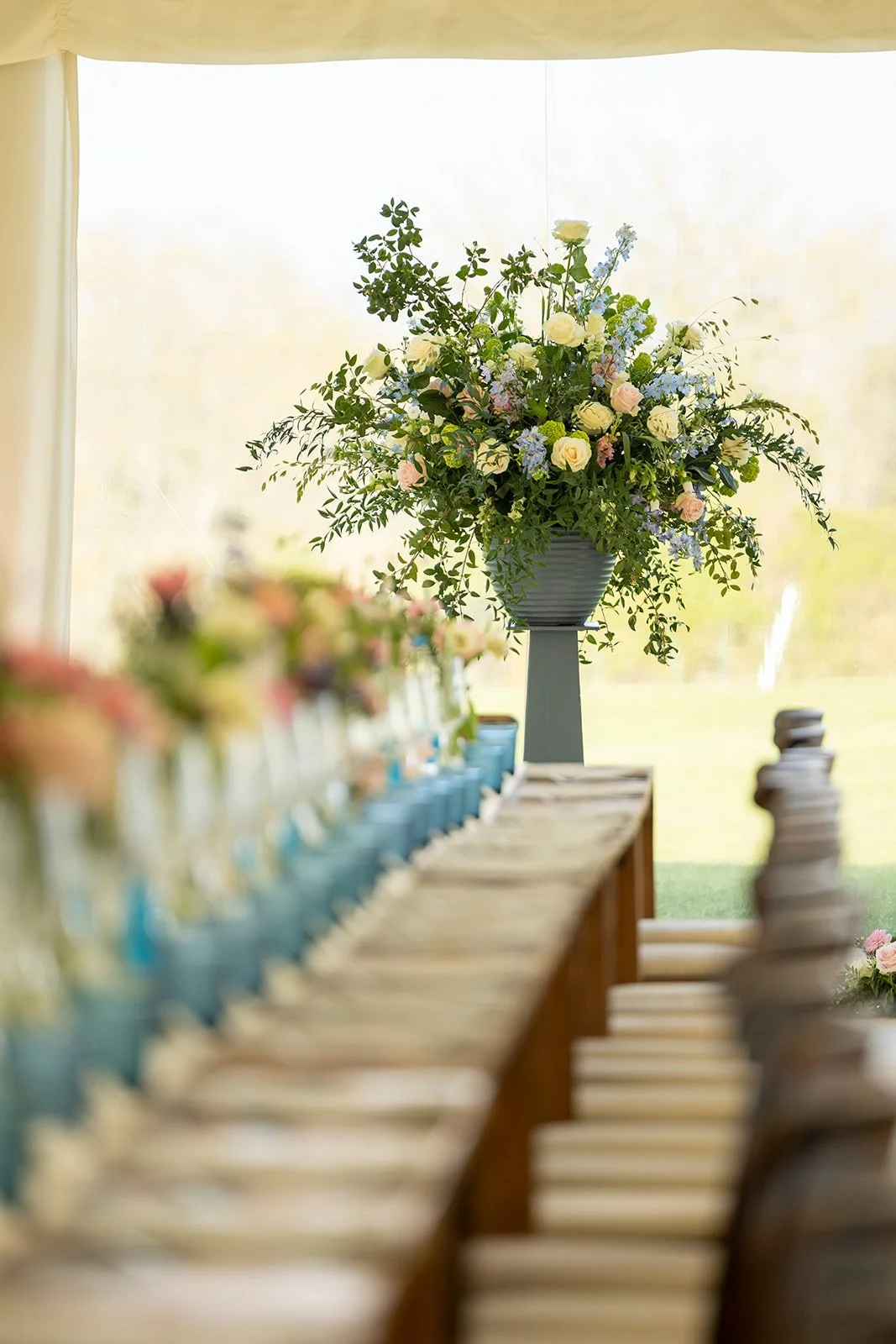 A large floral arrangement with white and pink roses, blue and green flowers, and greenery in a tall gray vase on a pedestal, with a table set with plates and glasses in the foreground, and a light-filled background.